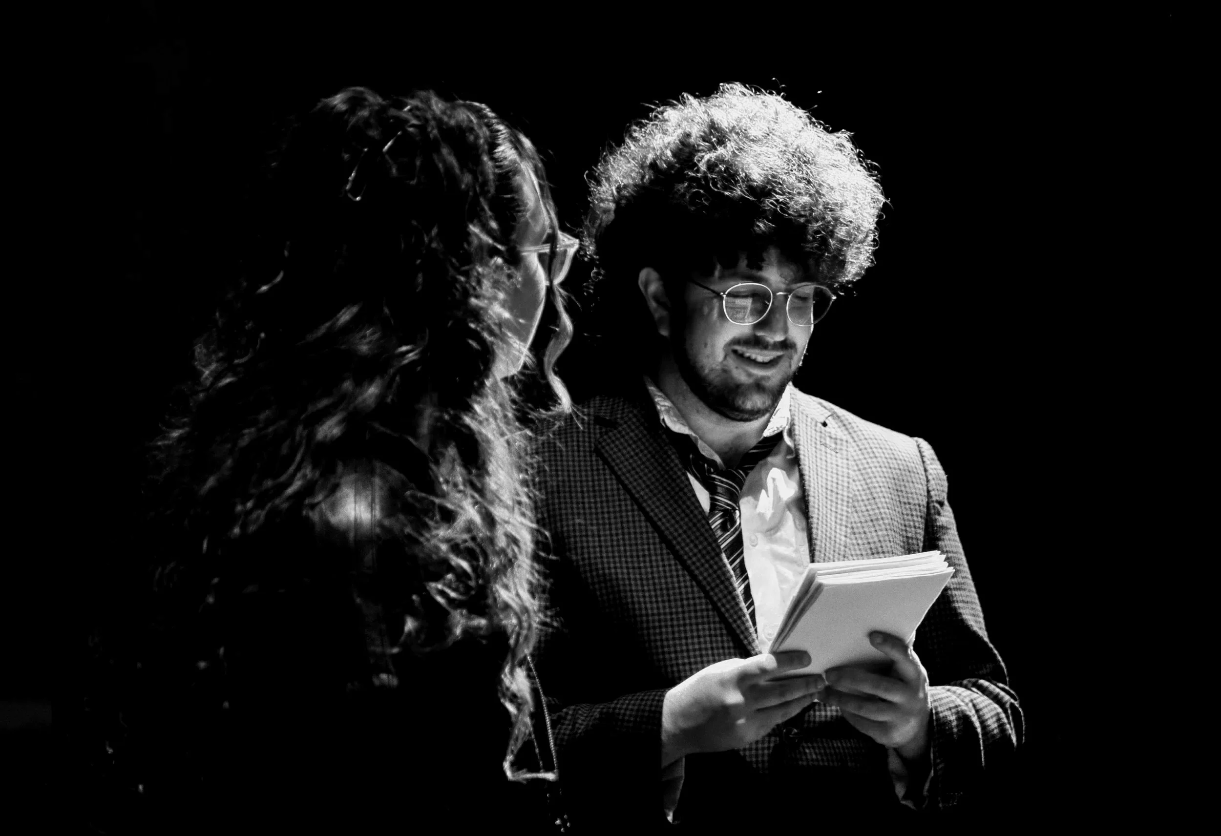 Two people, a woman with curly hair and glasses and a man with curly hair and glasses, are standing close together, looking at a book or document, in a dark environment with focused lighting.