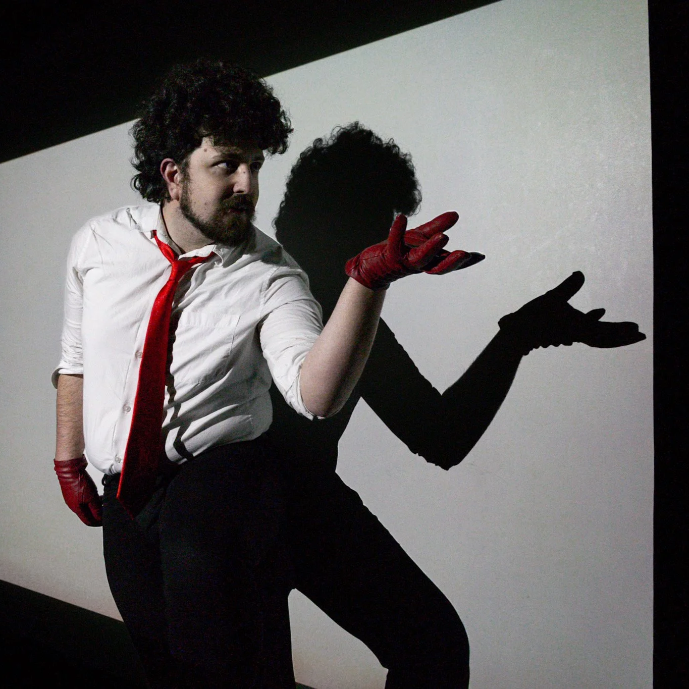 A man with curly hair and a beard wearing a white shirt, red tie, and red gloves, posing against a backdrop with his shadow projection creating a silhouette of a different pose.