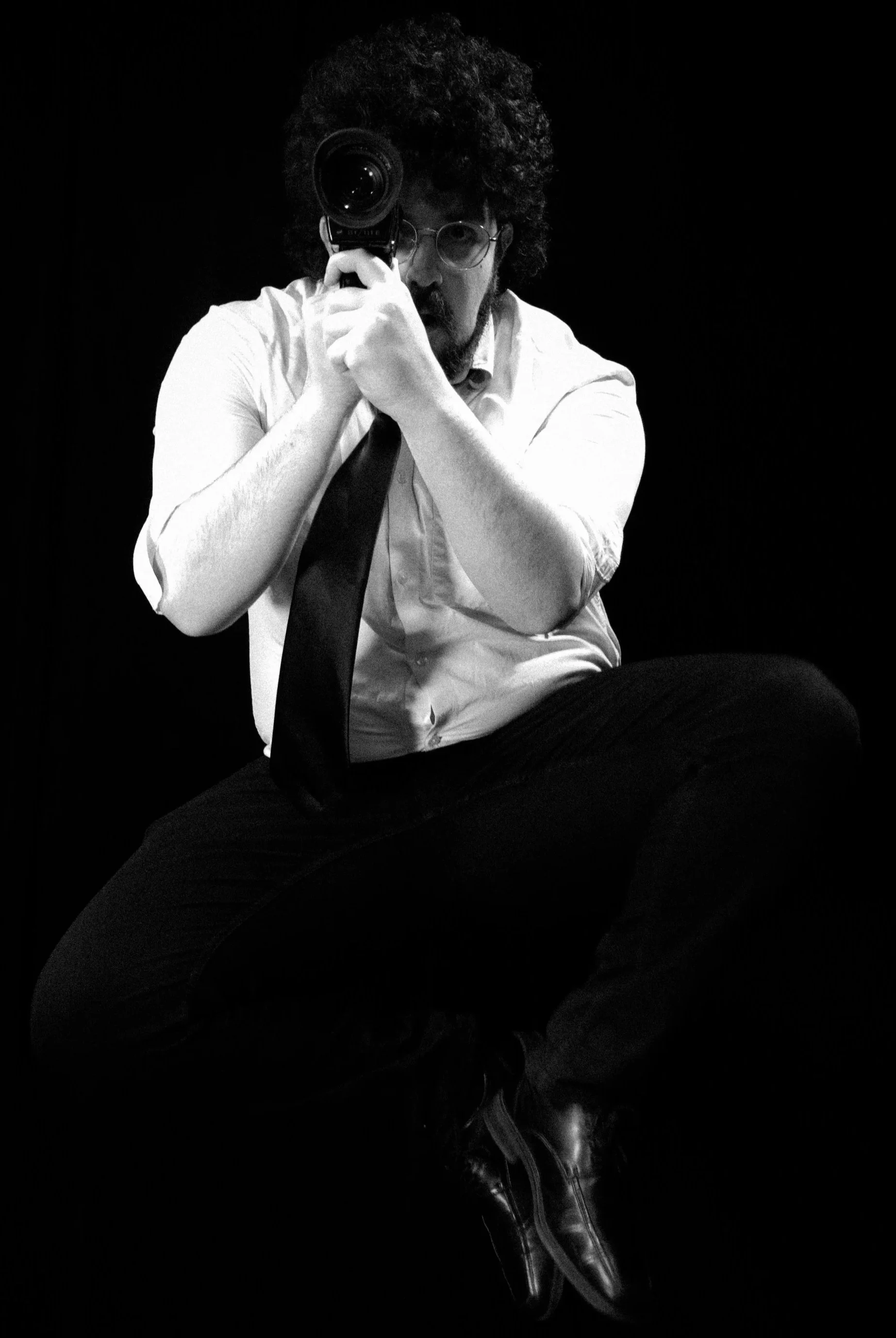 Black and white photo of a man with curly hair and glasses, sitting against a dark background, holding a camera lens to his eye, with his other hand supporting the camera.