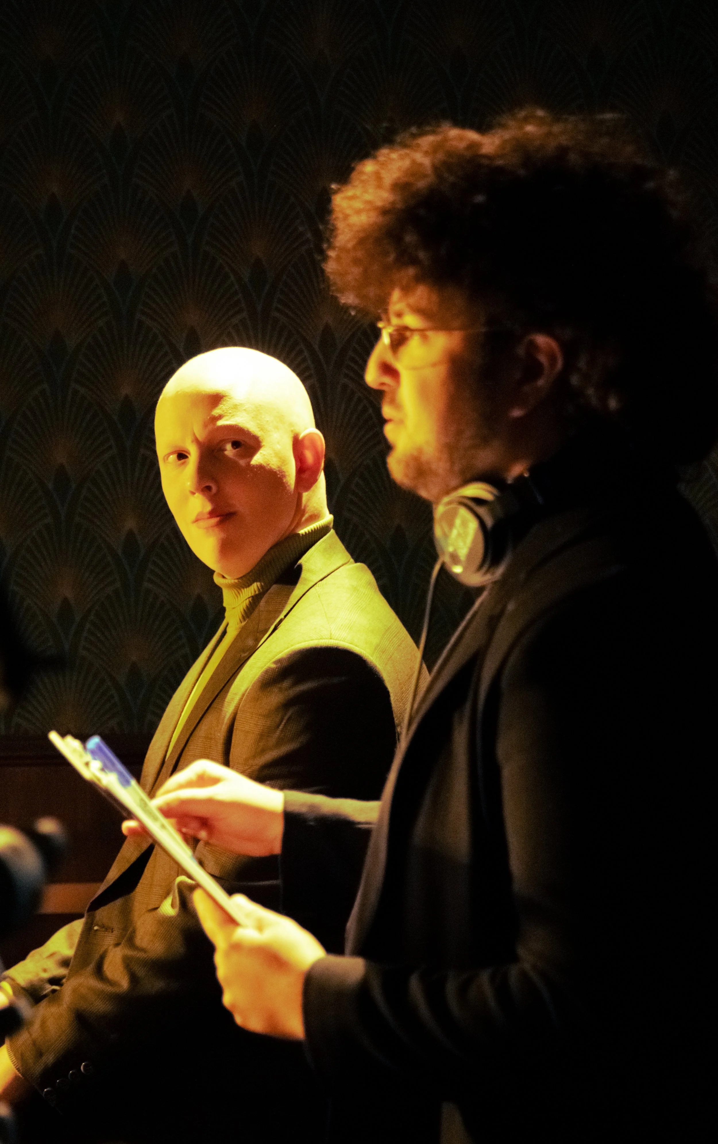 Two men in formal attire and glasses are seated indoors with patterned wallpaper in the background. The man on the left is bald and looking at the camera, while the man on the right has curly hair, beard, and headphones around his neck, and is looking downward, holding a smartphone.