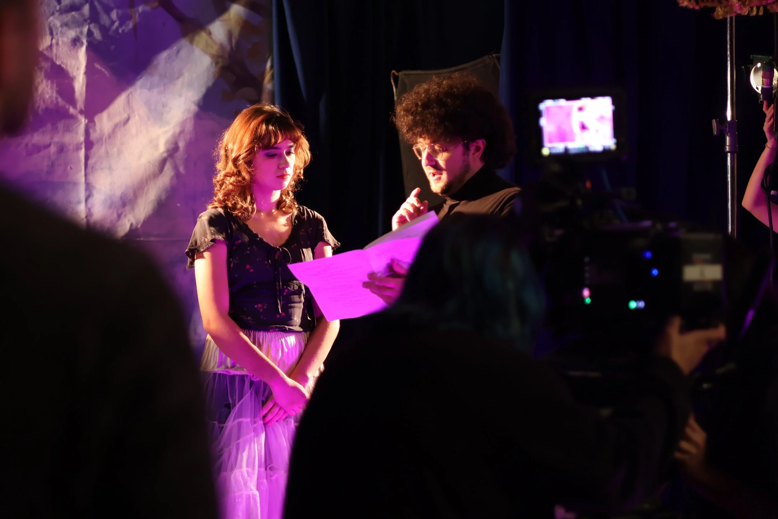 A young woman in a black floral top and purple tulle skirt is standing with a young man, possibly a director or scriptwriter, who is holding a script or notes. They are surrounded by filming equipment and are on a stage with a dark backdrop.