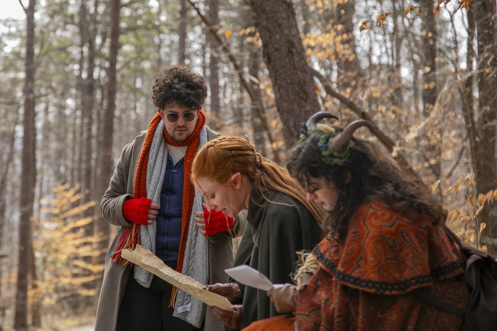 Three people in a forest, two women and one man, appear to be examining a map or document. One woman has red hair, the other wears animal horns and a colorful poncho. The man has curly hair, sunglasses, and is wearing a coat, scarf, and gloves. The background shows trees with autumn leaves.