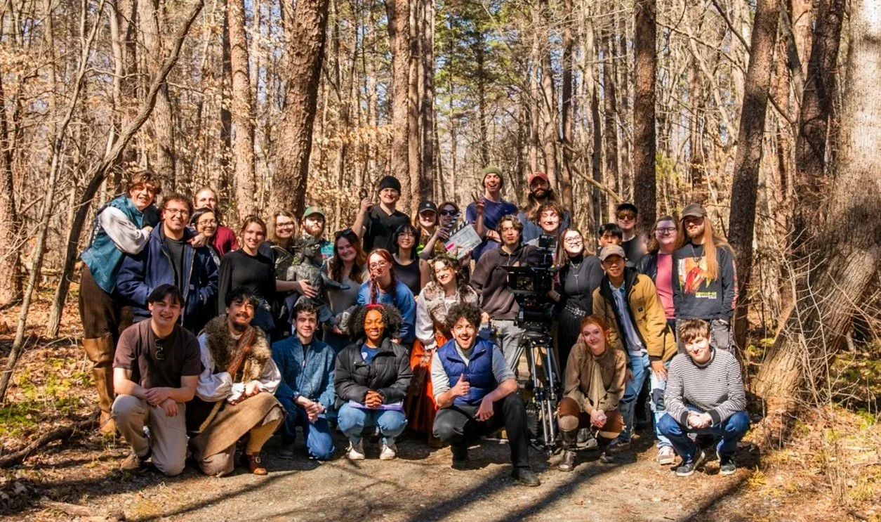 Group of people, including some with film equipment, standing in a wooded area with trees and sunlight.