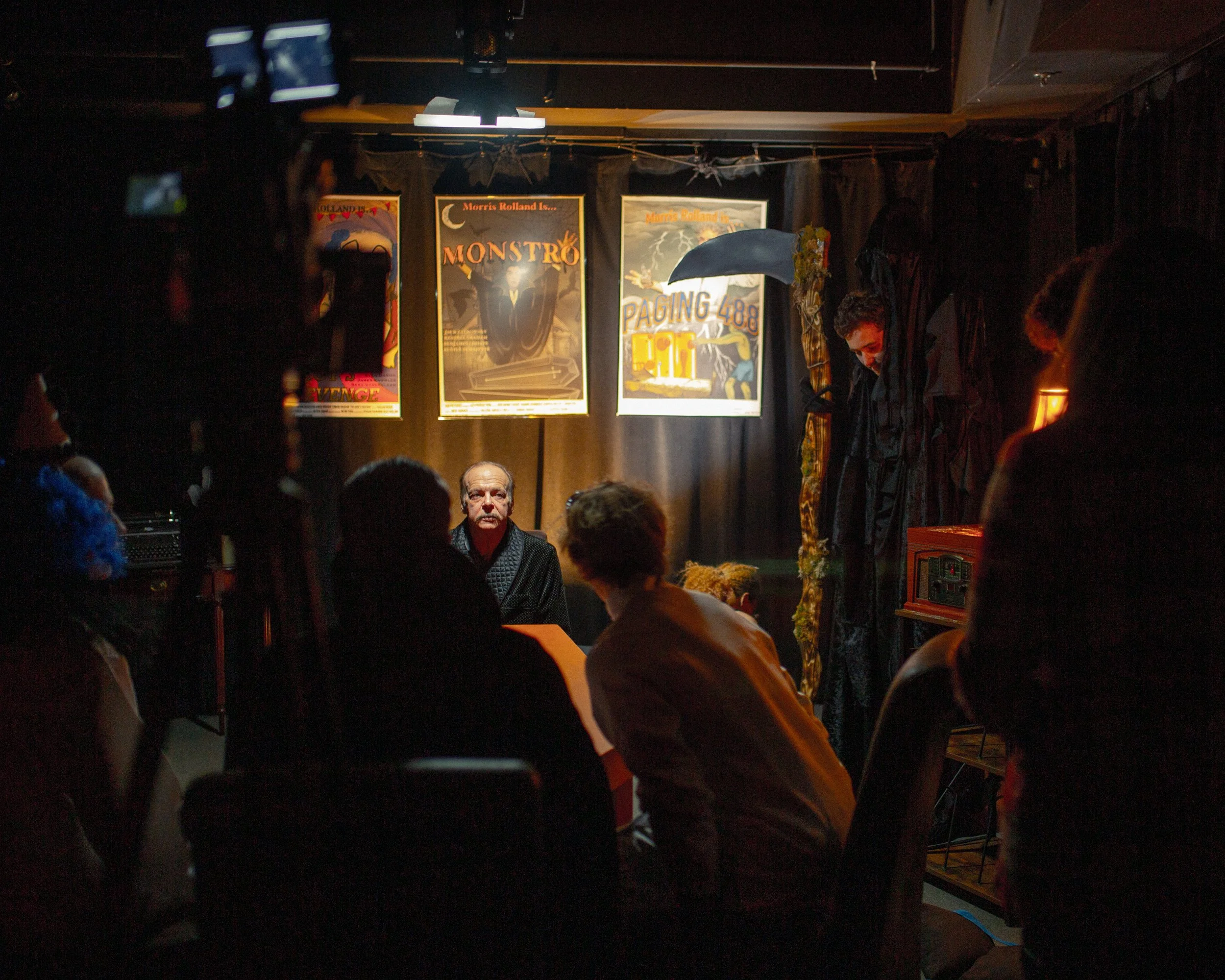 Stage with posters for horror movies, audience seated in front, one person on stage holding an umbrella, dark lighting.