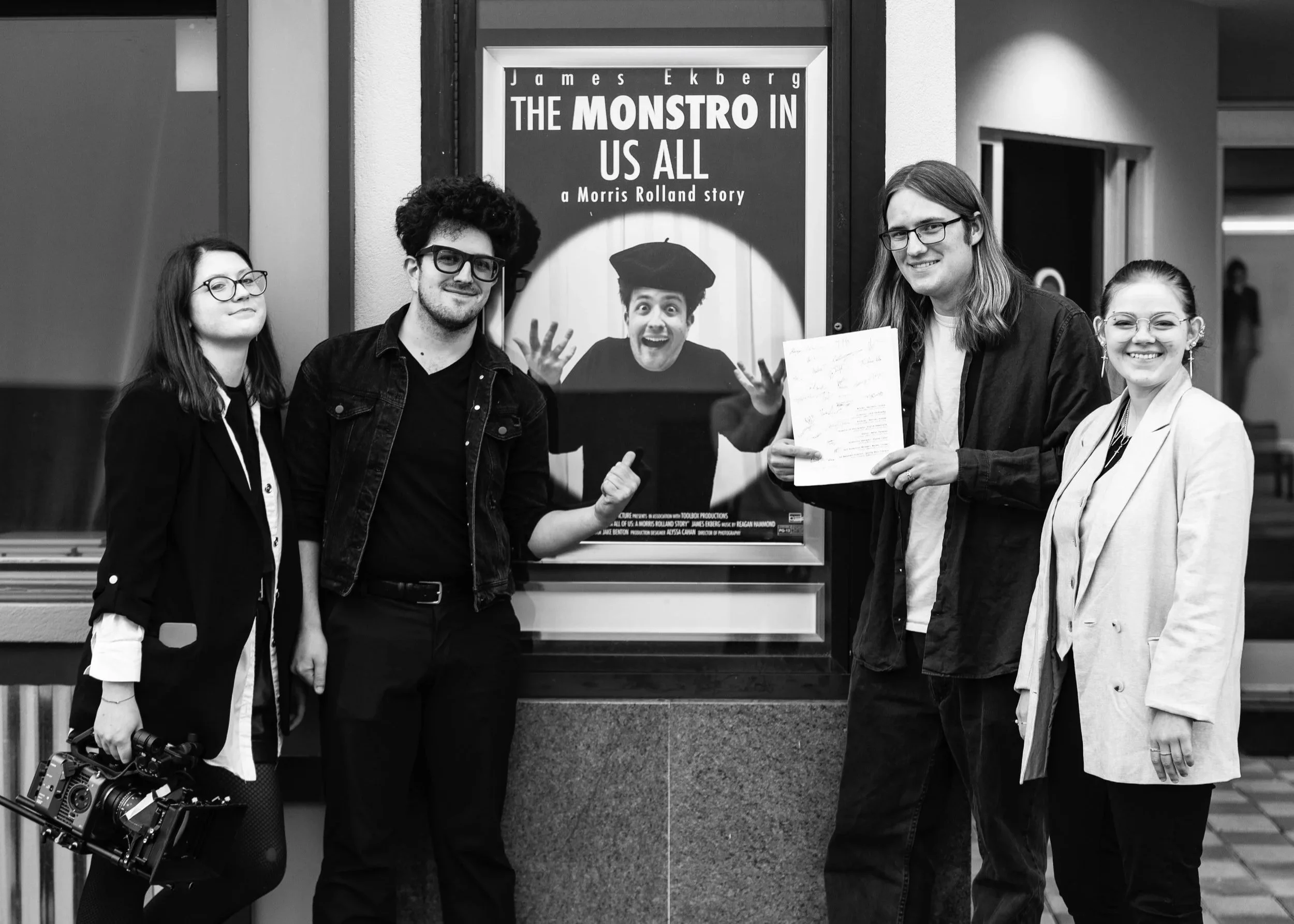 Four young adults standing in front of a movie poster for 'The Monstro in Us All,' smiling and posing for the photo. One person is holding a professional camera. The poster features a person with a large head wearing a graduation cap.