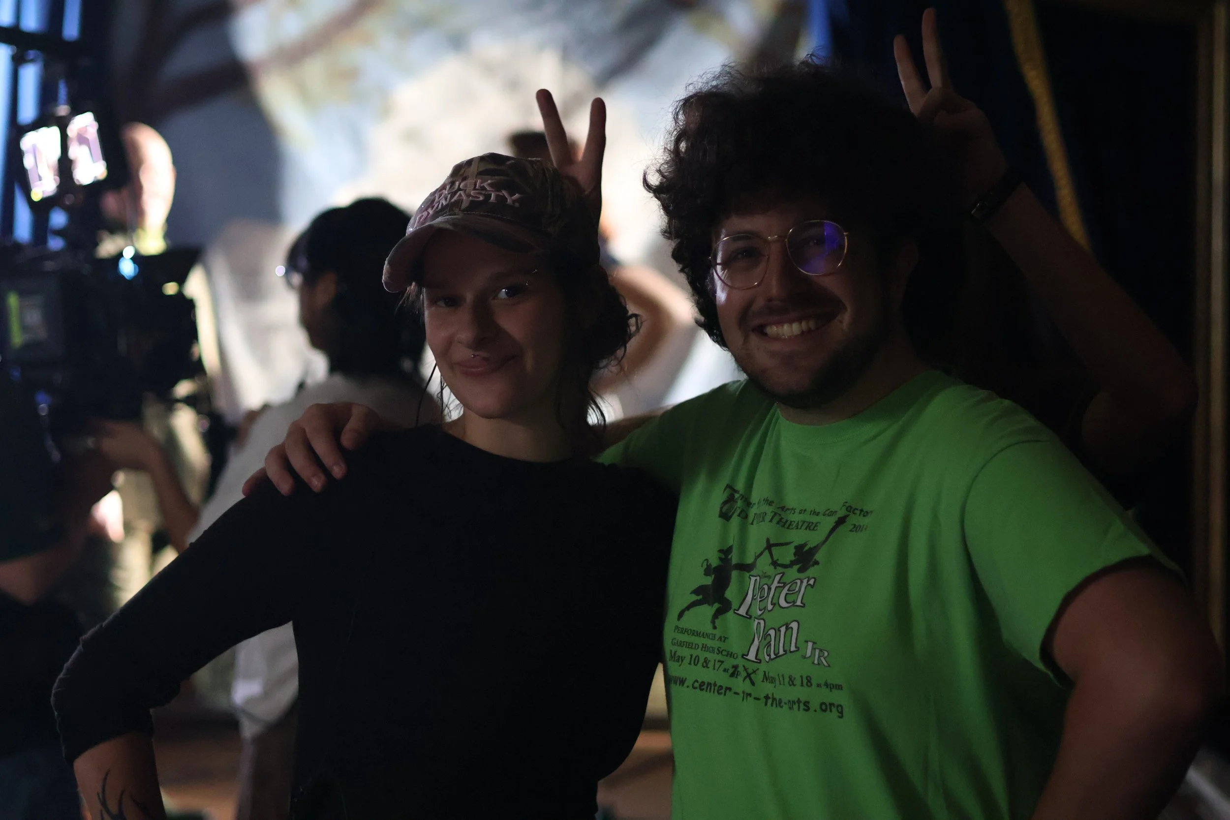 Two young people smiling at the camera, one making a peace sign behind the other. The person in front is wearing a bright green T-shirt with text on it. The scene appears to be indoors at a gathering or event.