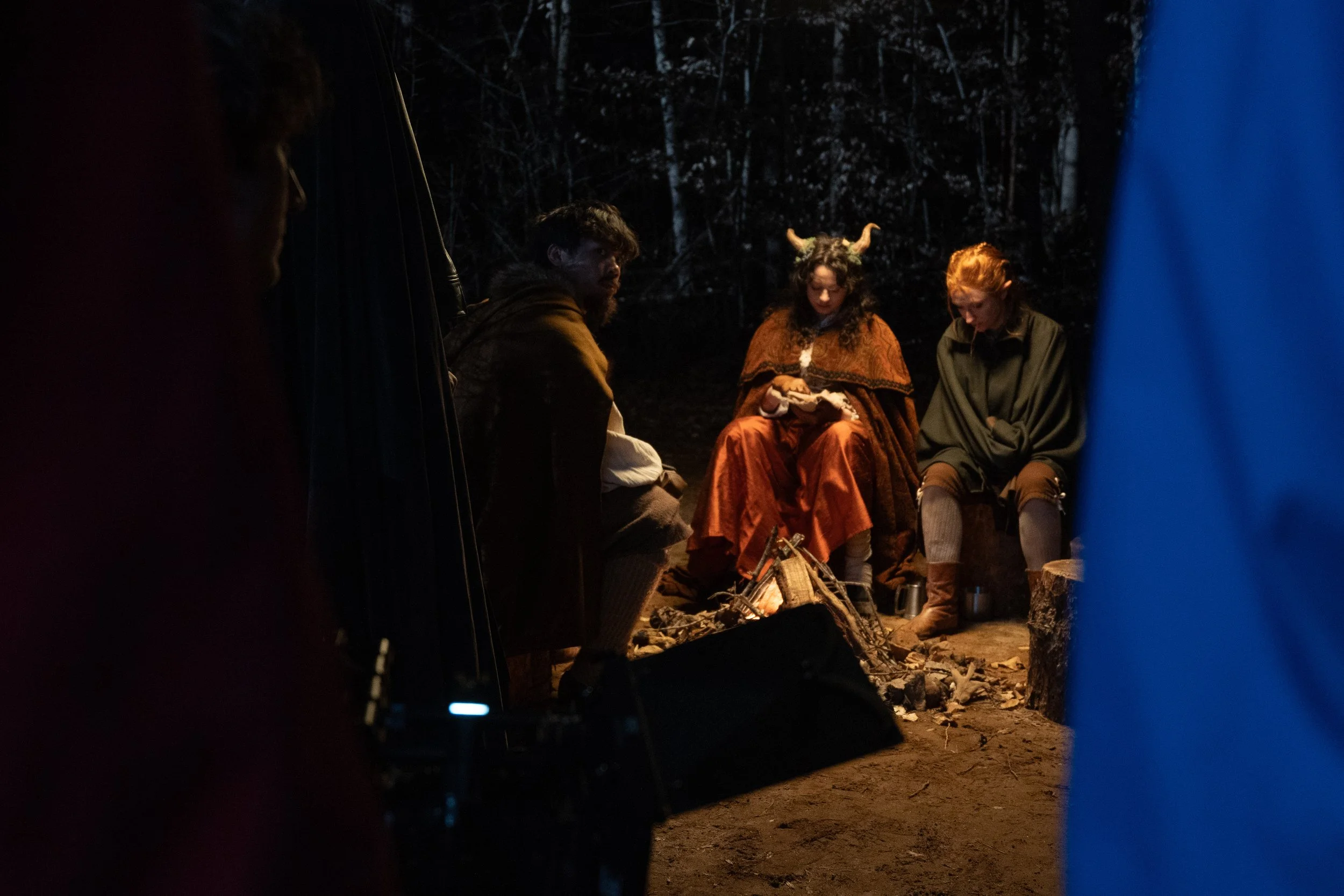 A group of people sitting around a campfire outdoors at night, with one person wearing horns on their head and dressed in a costume.