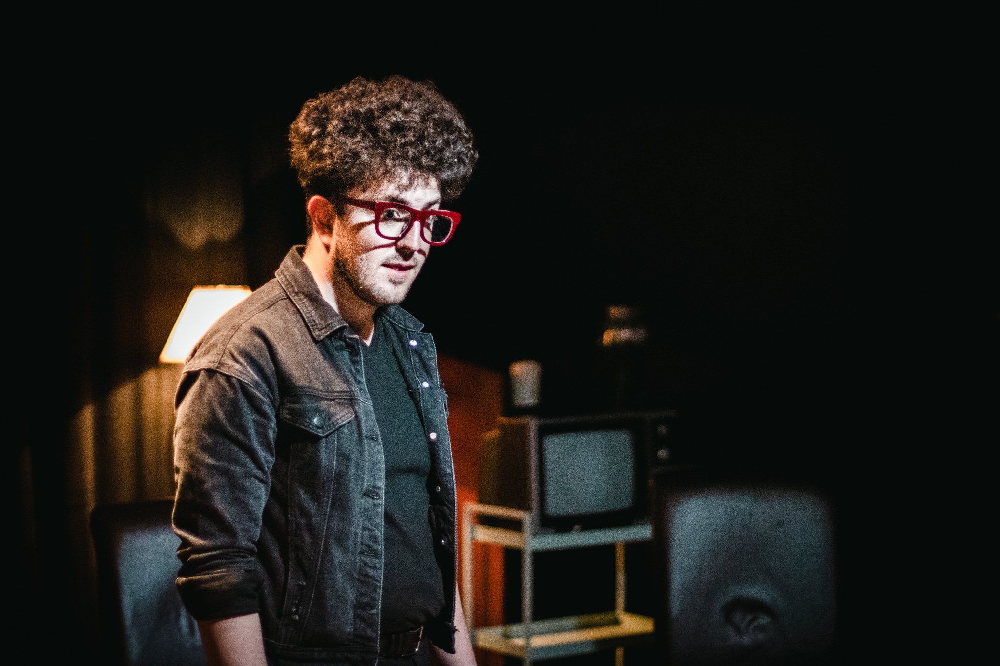 A person with curly hair wearing red glasses and a dark denim jacket, standing in a dimly lit room.