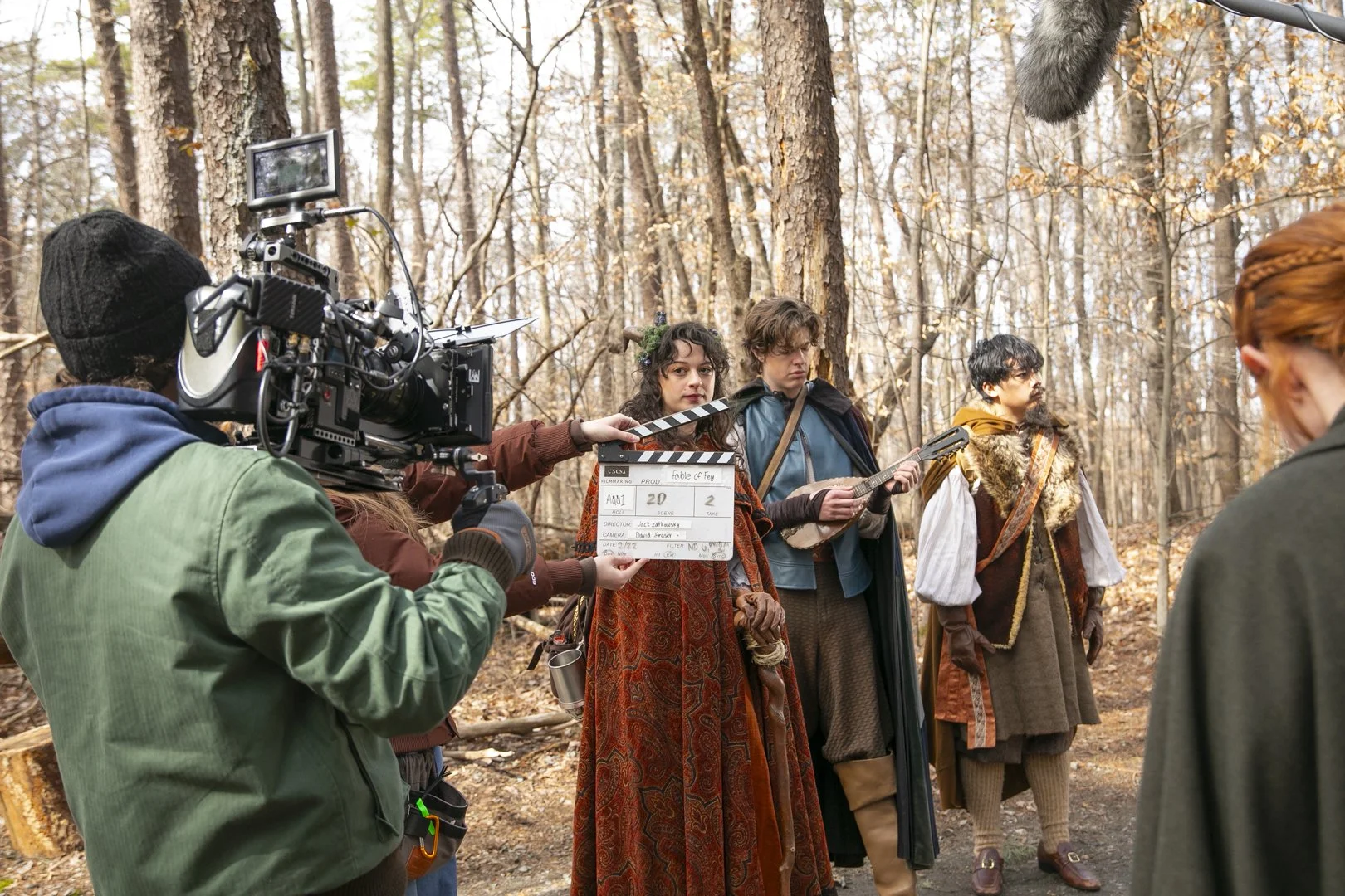 Filming of a movie scene set in a forest with actors in fantasy costumes, crew members, and equipment including a camera and clapperboard.
