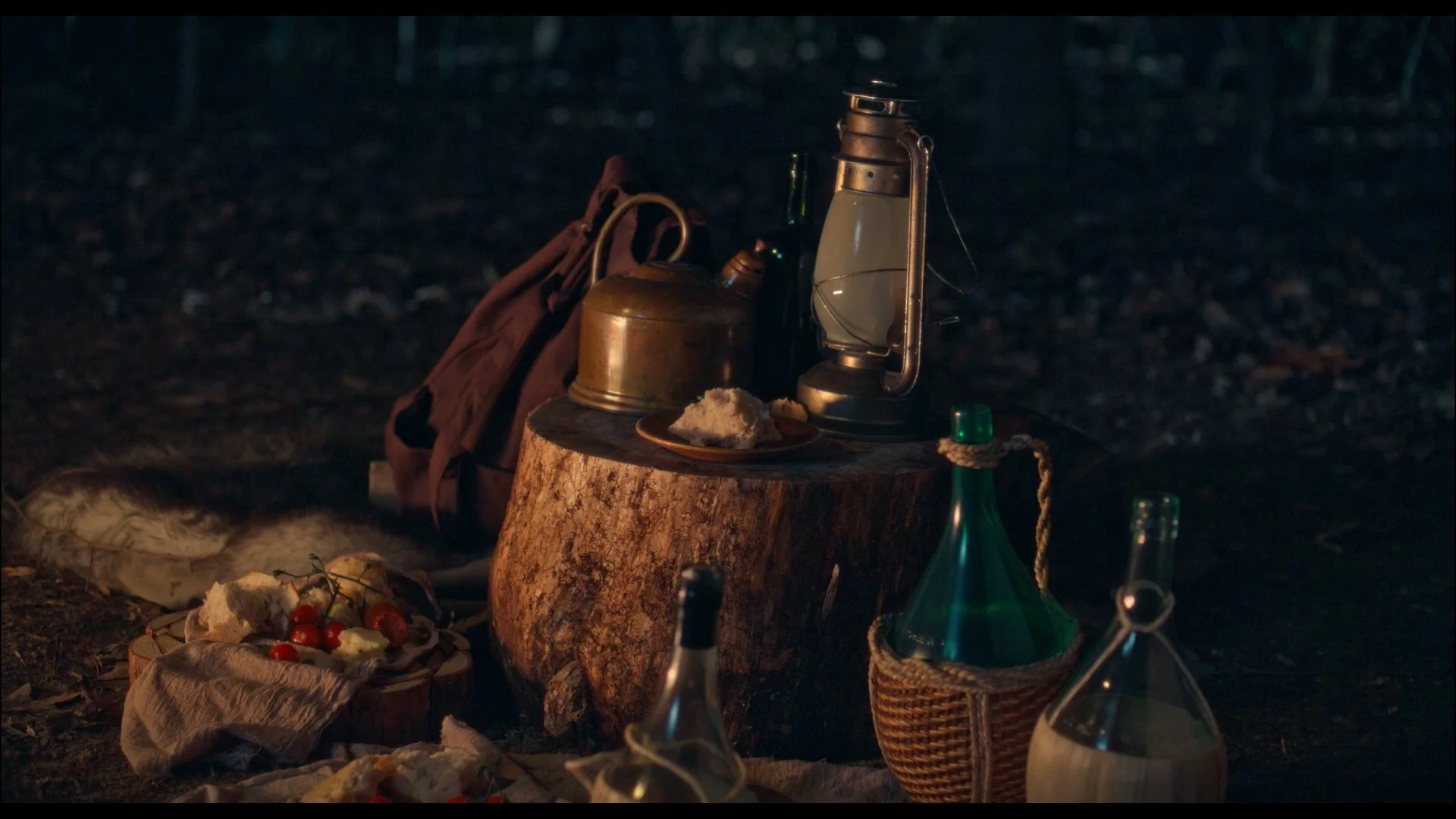 Campfire setup in a dark forest with vintage lanterns, bottles, a wooden stump, a plate of bread, cherry tomatoes, and other food items on blankets.