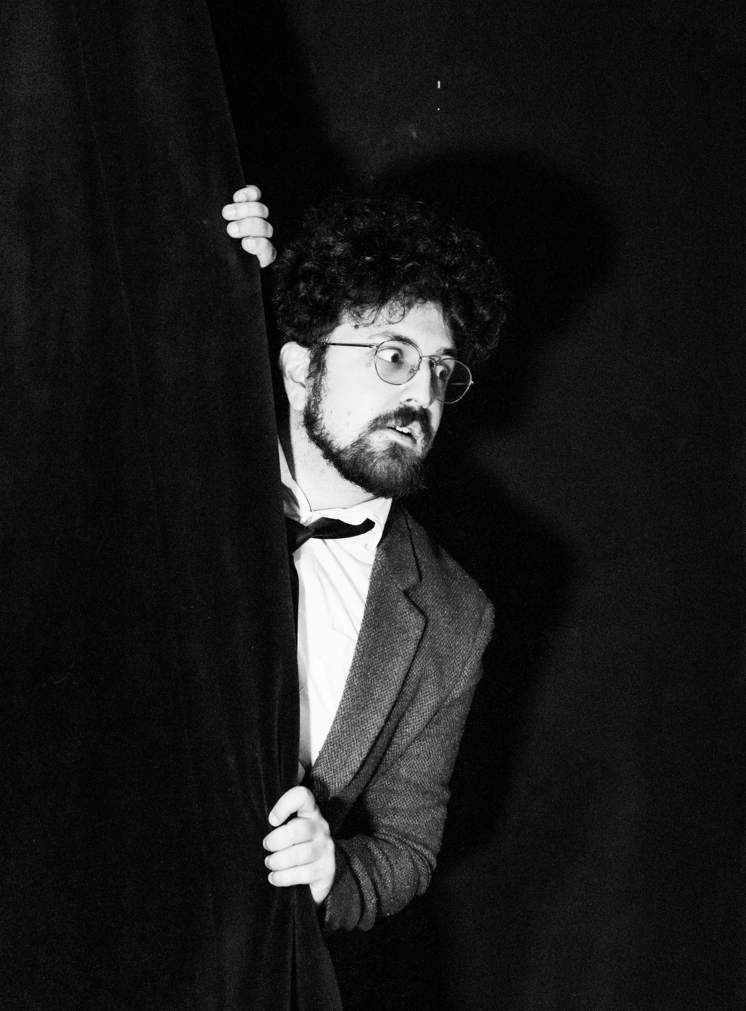 Black and white photo of a man with curly hair, beard, and glasses, partially hidden behind a dark curtain or wall, looking to the right.