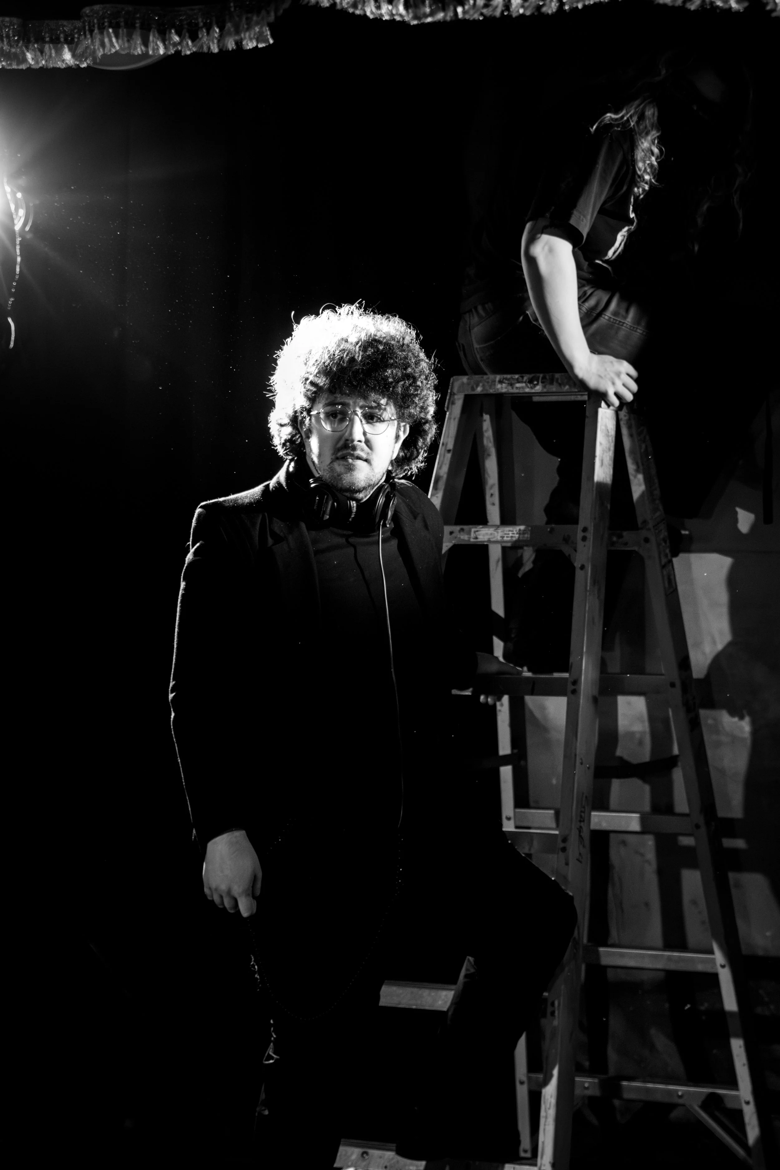 Black and white photo of a man with curly hair, glasses, and headphones around his neck, standing in front of a dark background. A woman is in the background on a ladder, facing away, with hair covering her face and hand gripping the ladder. There is a bright light source on the left side.