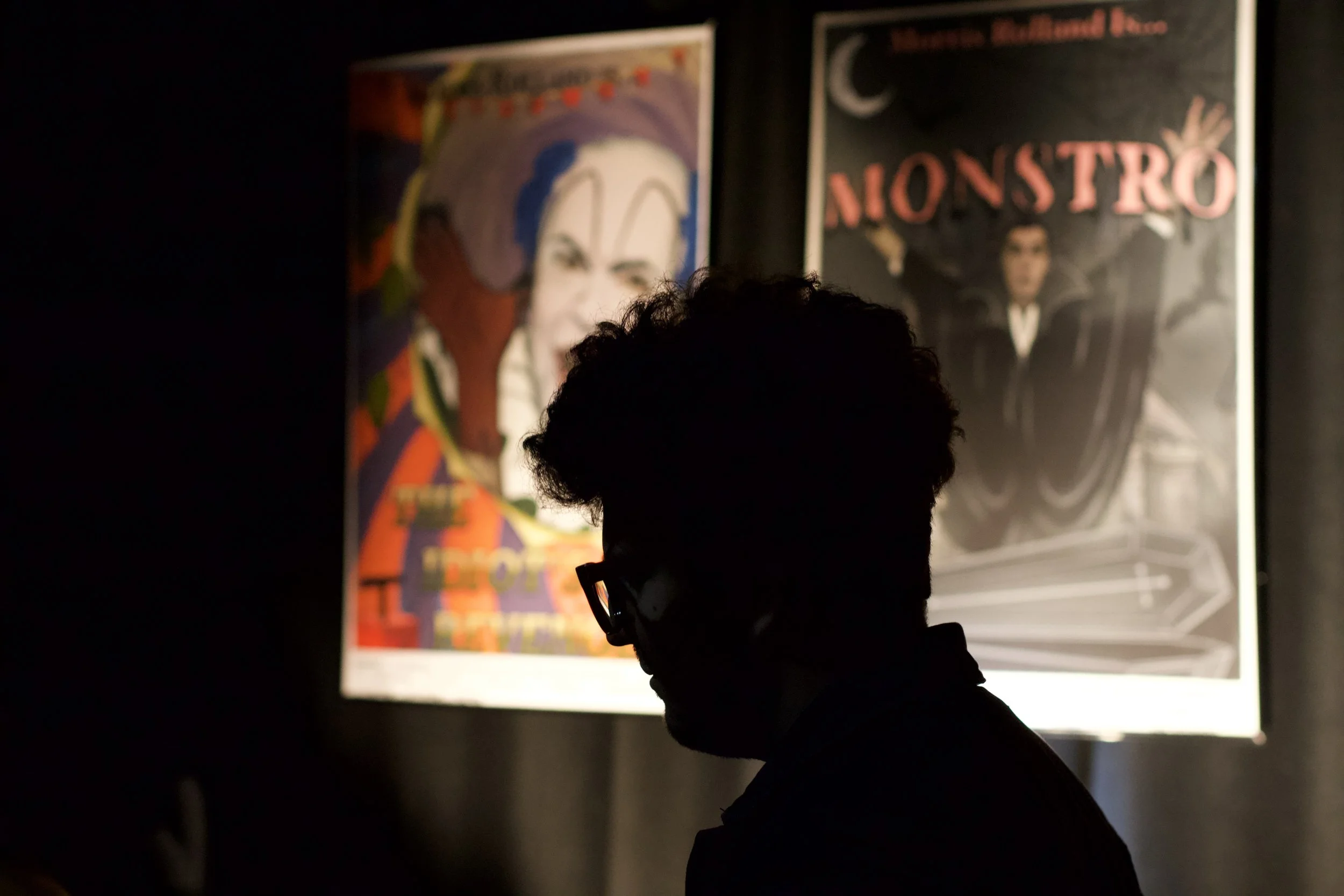 Silhouette of a person with curly hair and glasses looking at illuminated movie posters on a dark wall.
