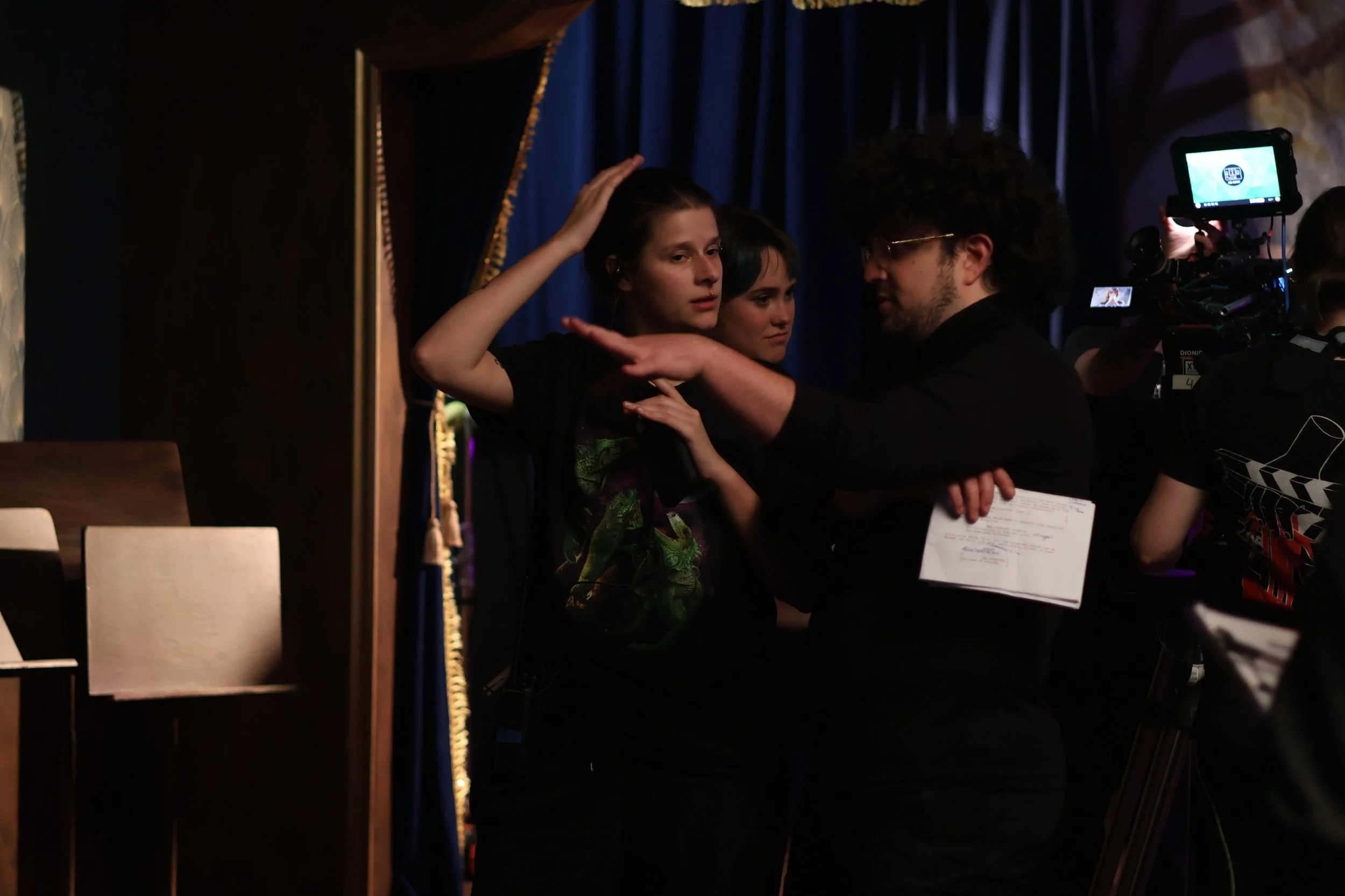 A film director instructs a female actress on set, with a camera operator filming nearby and dark blue curtains in the background.