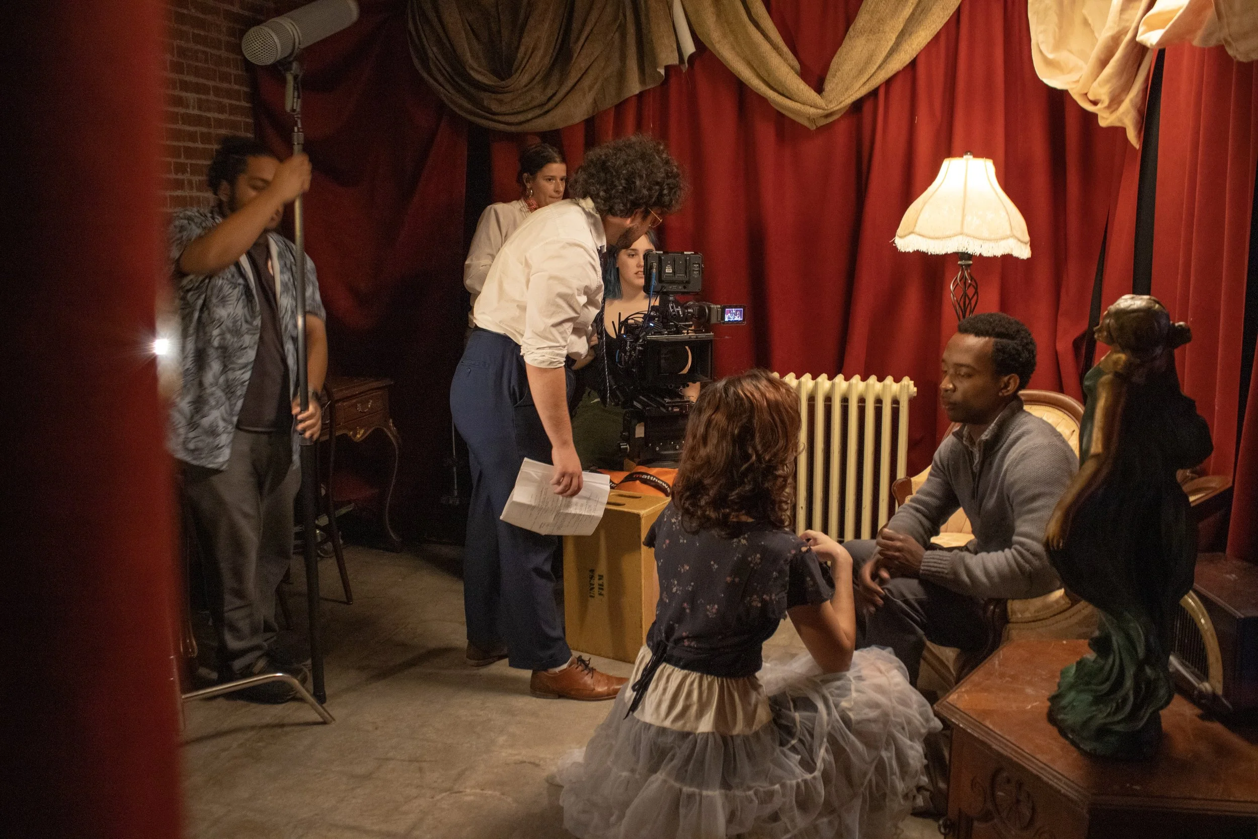 A behind-the-scenes look at a film or video production crew setting up a scene in a cozy, dimly lit room with red curtains. Several people are gathered around, some sitting and some standing, with one person operating a camera. There is a vintage-sty