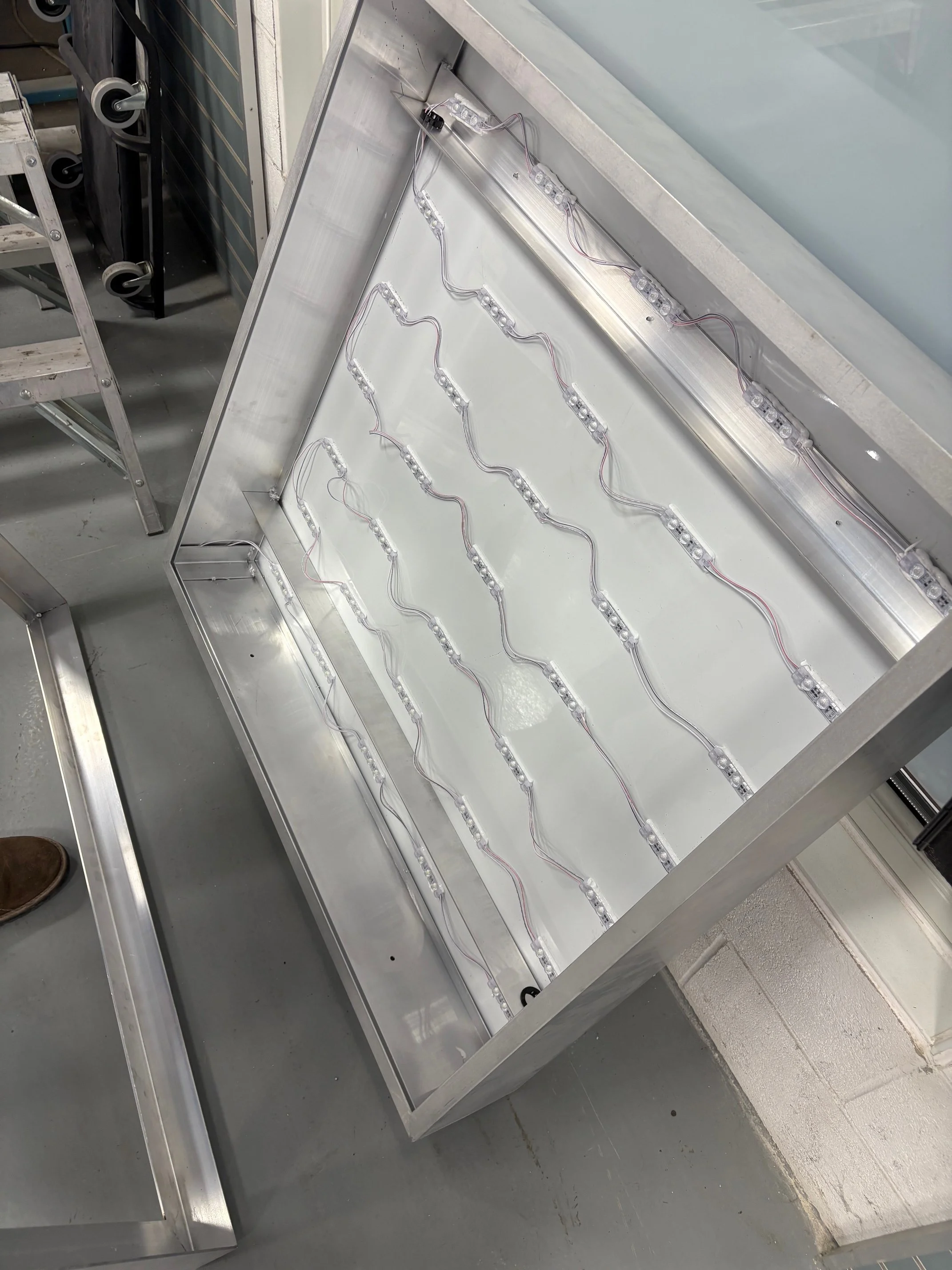 An indoor workspace showing a metal fabricated sign frame or box with several rows of LED strip lights connected by wires, this newly built custom sign cabinet is in the process of assembly.