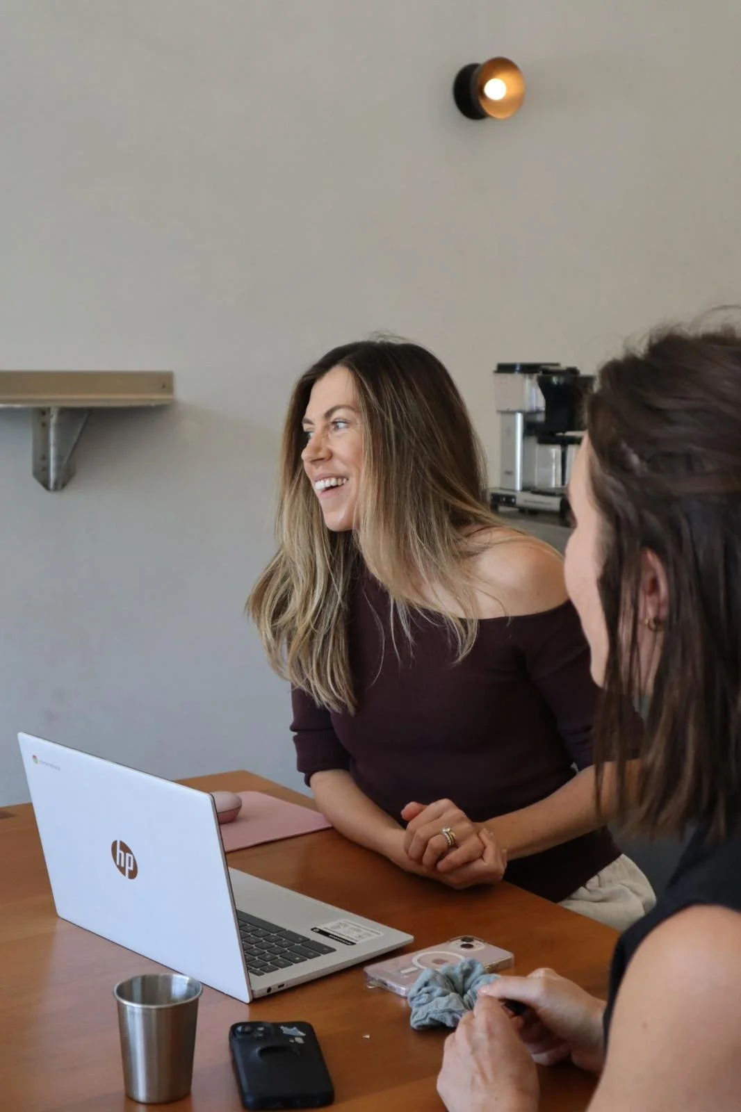 Two women sitting at a wooden table with a white HP laptop, one woman with long brown hair smiling and looking to the side, the other with short dark hair holding a phone and a scrunchie, a metal cup, and a smartphone on the table, in a room with a shelf and a coffee machine in the background.