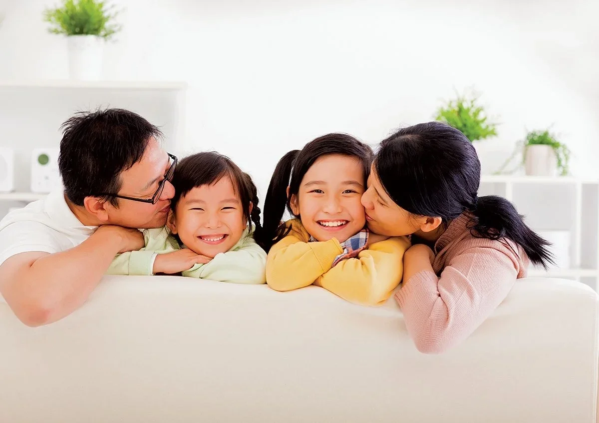 Family of four with two children, sitting on a white couch, smiling, hugging, and showing affection in a bright living room with white walls and green indoor plants.