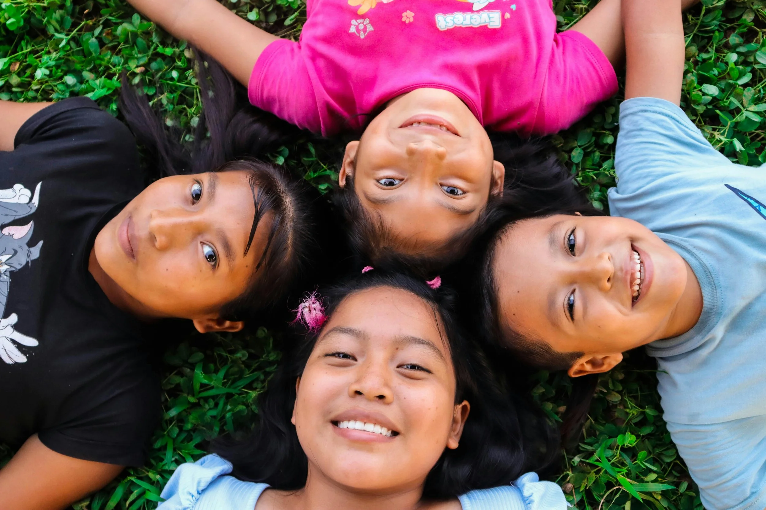 Five children lying on green grass, smiling and looking up at the camera.