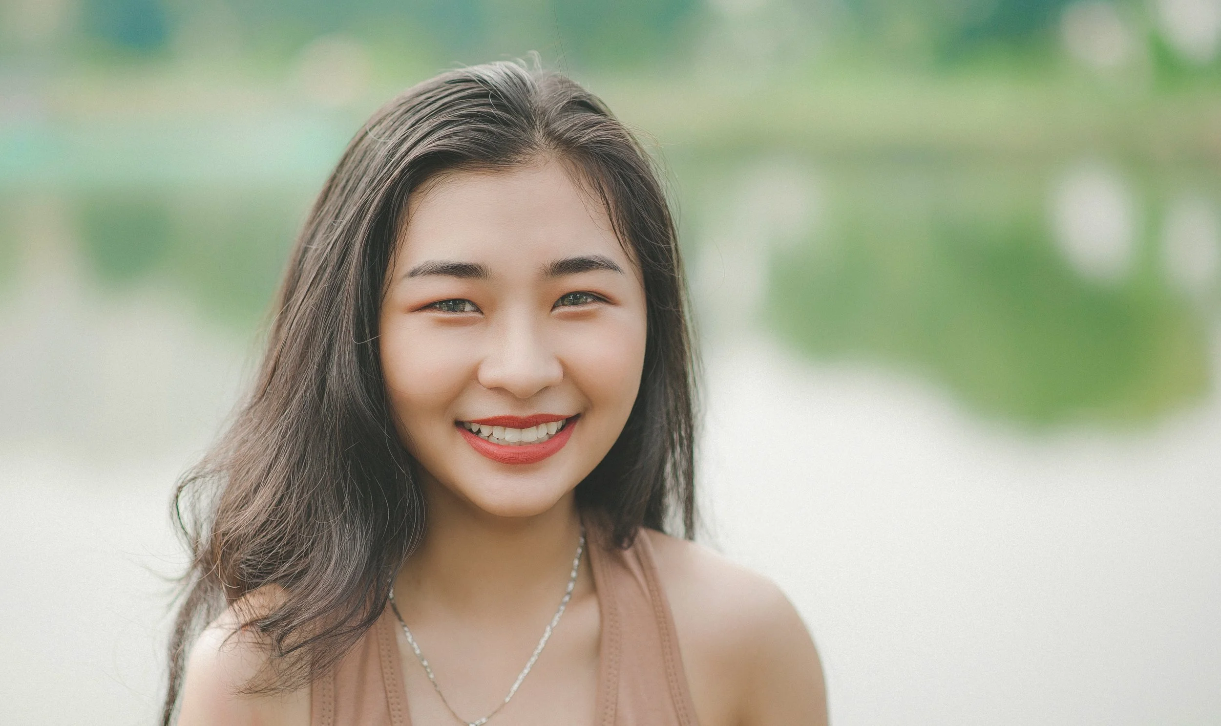 A young woman with dark hair, smiling, wearing a beige sleeveless top and jewelry, outdoors with a blurred lush green background.