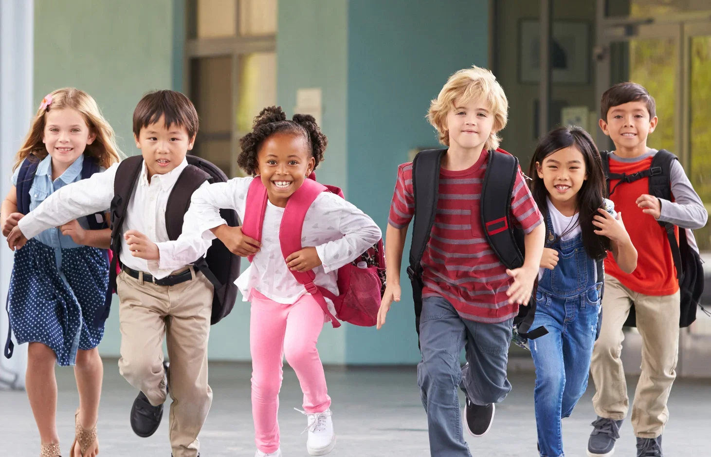 A group of six diverse children running and smiling, carrying backpacks, outside of a school building.