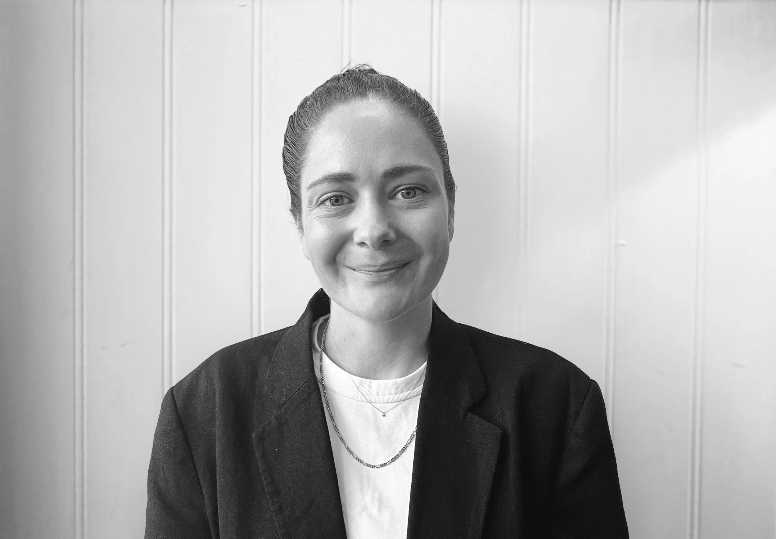 Black and white portrait of a woman with hair tied back, smiling, wearing a black blazer and layered necklaces, posed in front of a paneled wall.