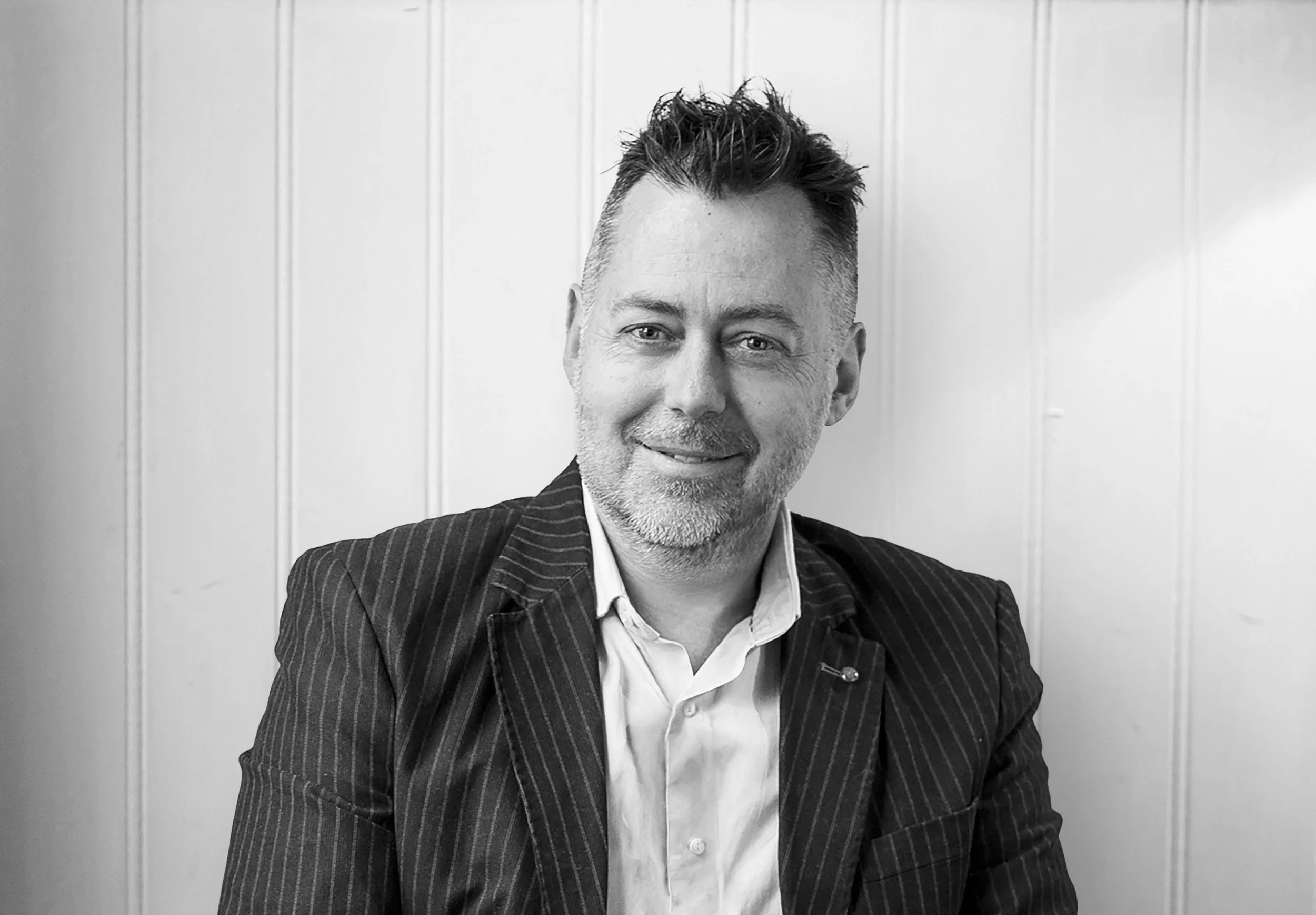 Black and white portrait of a middle-aged man with short, spiked hair, a beard, wearing a pinstripe suit and white shirt, sitting against a paneled wall background.