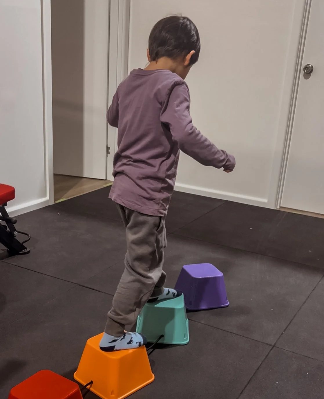 A young boy with dark hair, wearing a mauve long-sleeved shirt, grey pants, and socks, is balancing on upside-down colored buckets in a room with white walls and a dark floor.