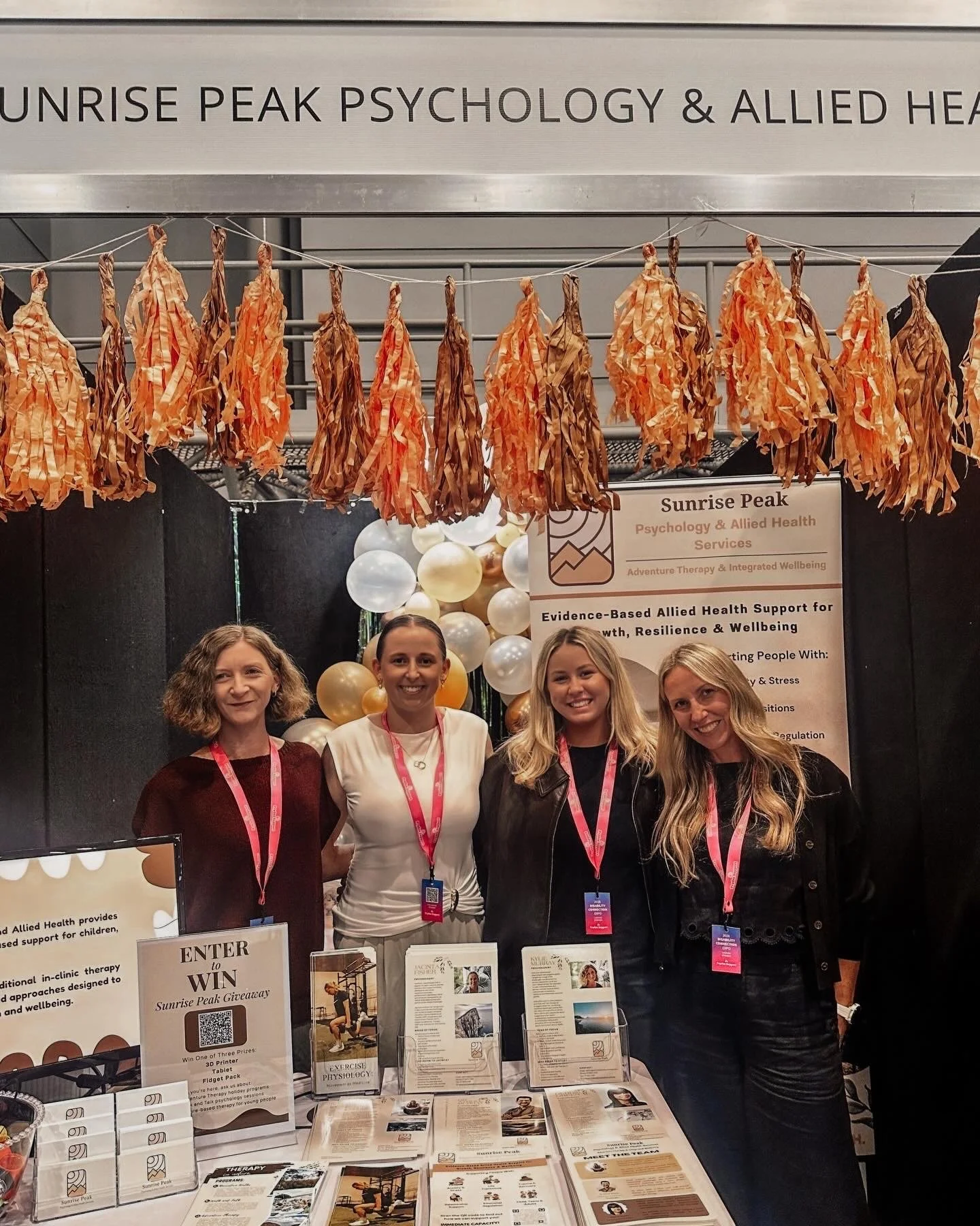 Team members from Sunrise Peak Psychology and Allied Health Services at an event booth with promotional materials and signage