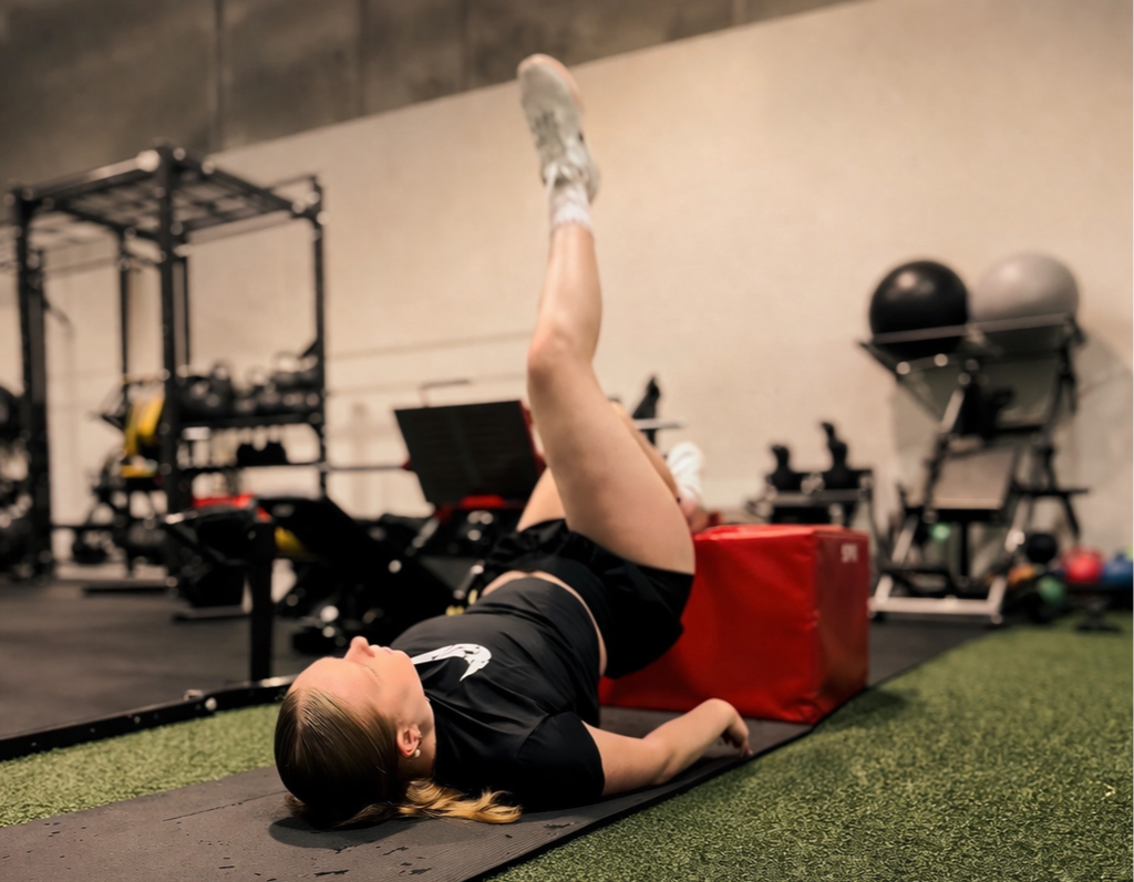 Woman performing a single leg glute bridge on a mat with a box in a gym, focusing on strength and movement control