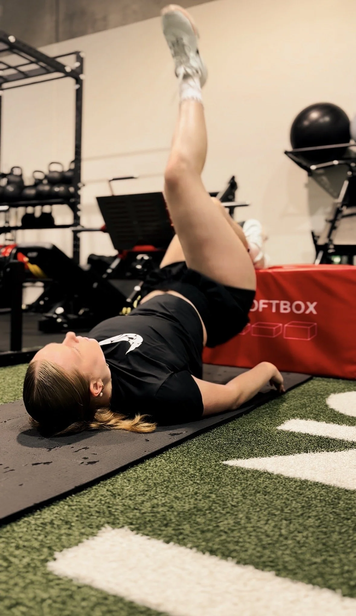 Woman performing a single leg glute bridge on a mat with a box in a gym, focusing on strength and movement control