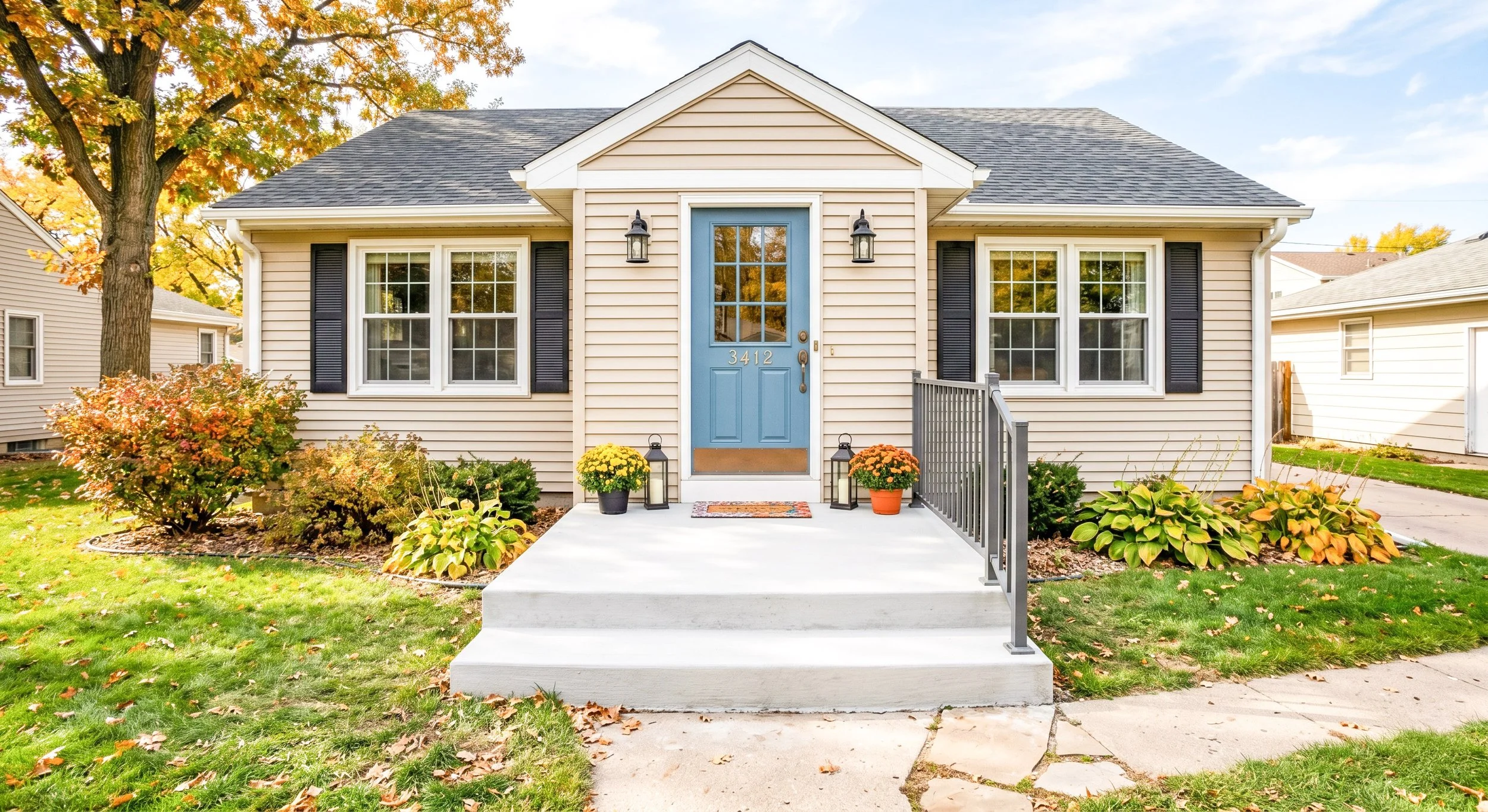 Front view of a beige house with a blue front door, black shutters, and two black outdoor lanterns. There are plants and autumn leaves in the yard, with a sidewalk and stairs leading to the entrance.