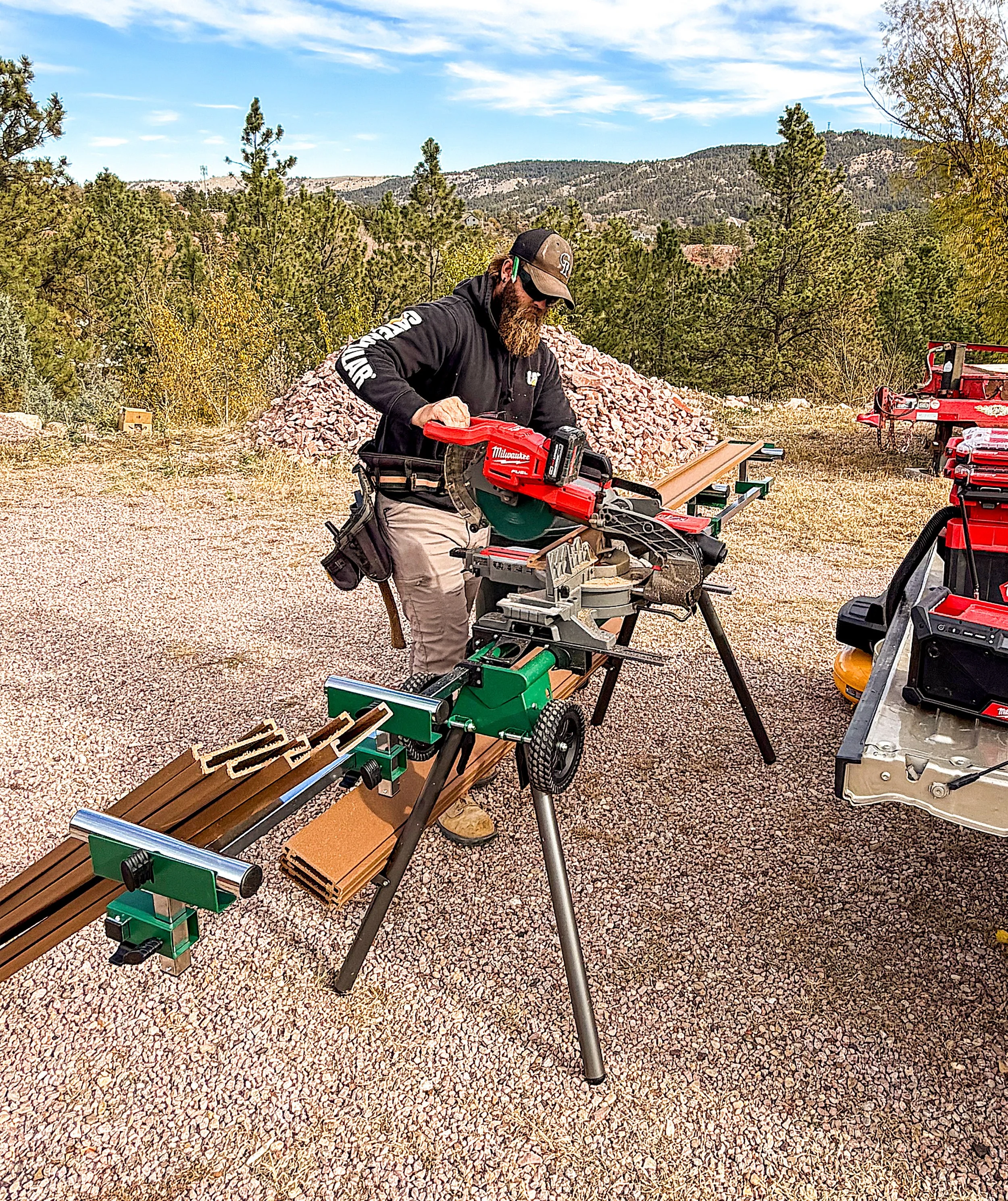 Man working with a miter saw outdoors in a mountainous area with trees and rocks, wearing a black hoodie, cap, and sunglasses.