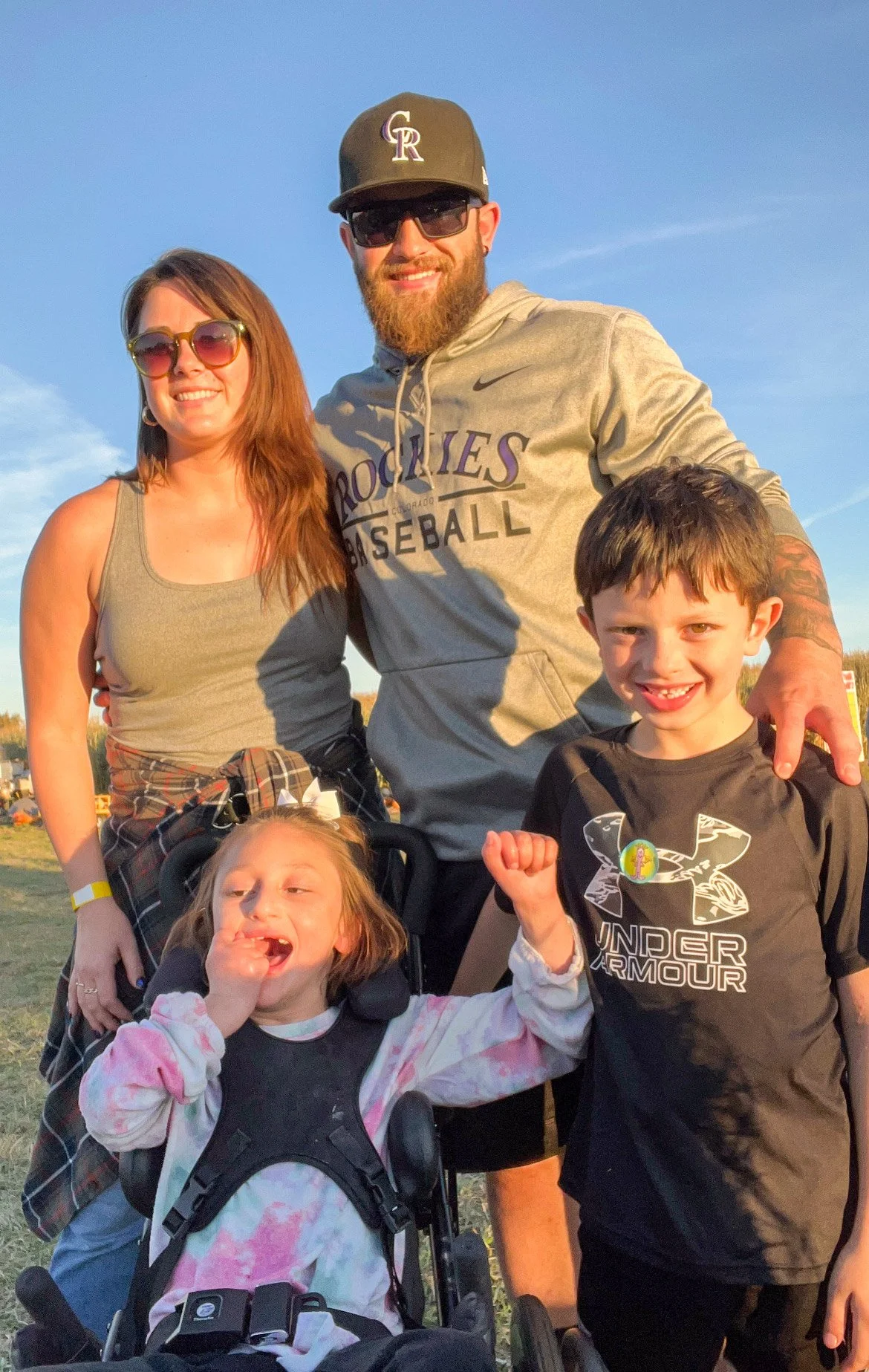 A group of four people, including two children and two adults, outdoors during the daytime. All are smiling and appear happy. The man is wearing sunglasses, a Colorado Rockies hoodie, and a cap, while the woman is wearing sunglasses and a casual tank top. One child is in a stroller, wearing a tie-dye shirt, and making a peace sign, while the other child is standing, wearing a black Under Armour shirt, and showing a fist.