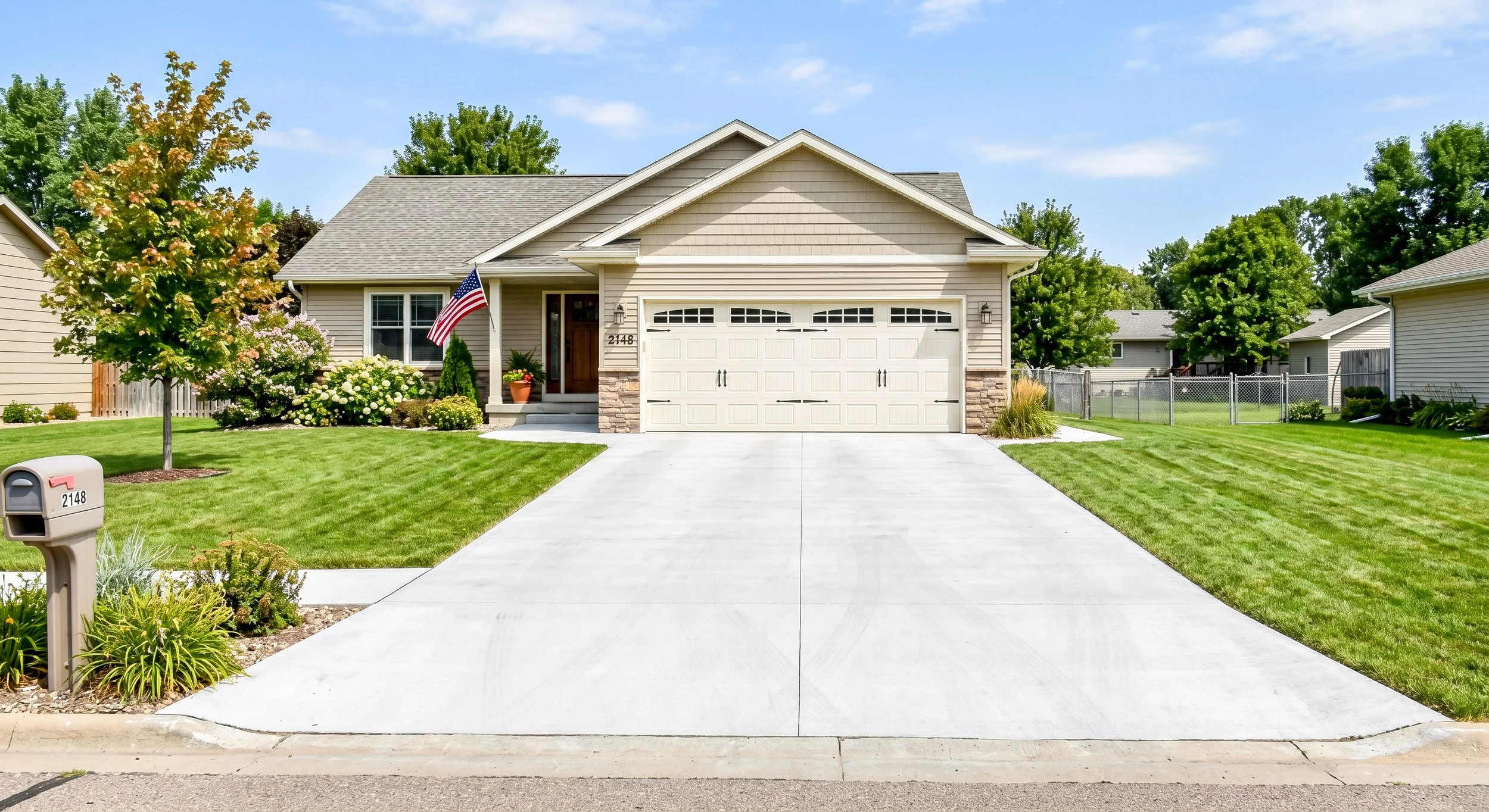 A suburban house with a well-maintained front yard featuring green grass, trees, and flower bushes. The house has beige siding, a double garage door, and a flag hanging near the entrance. A mailbox with the number 2148 is visible in the yard.