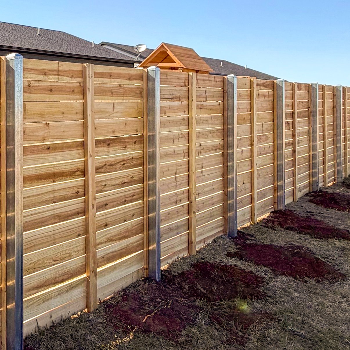 A newly constructed wooden privacy fence with vertical wooden panels and metal posts in a backyard, on a dirt ground under a clear blue sky.