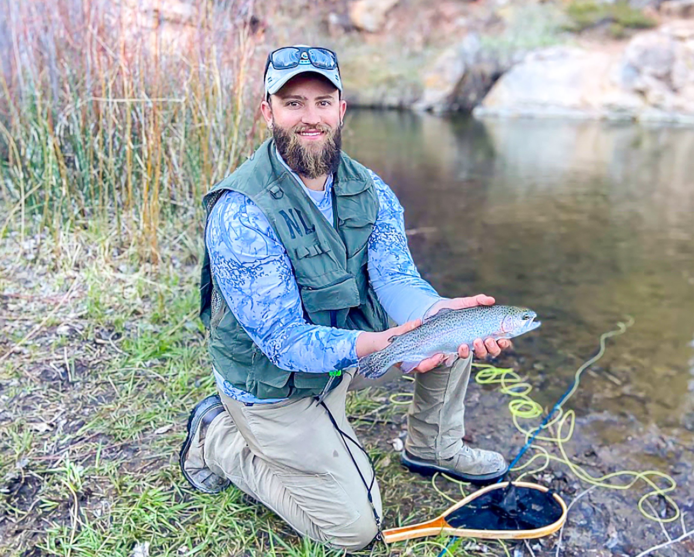 A man with a beard, wearing a cap, sunglasses, and outdoor gear, kneeling by a river bank and holding a large fish he caught.