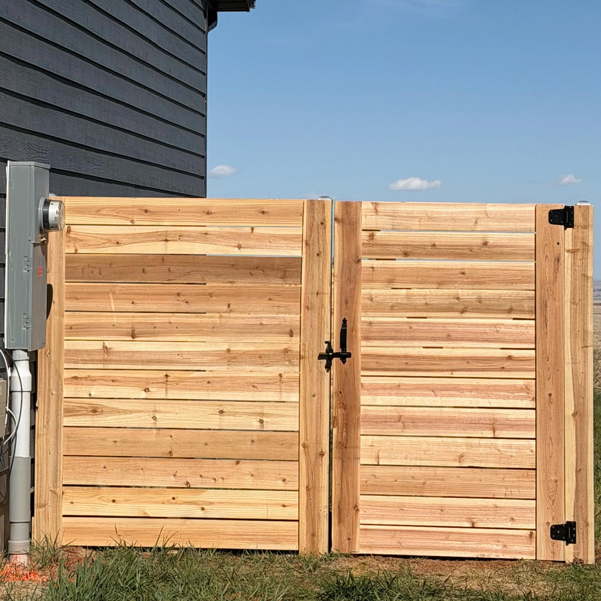 A wooden privacy fence with a black metal latch, partially enclosing a yard with grass, beside a gray house exterior with horizontal siding, and a blue sky with small clouds in the background.