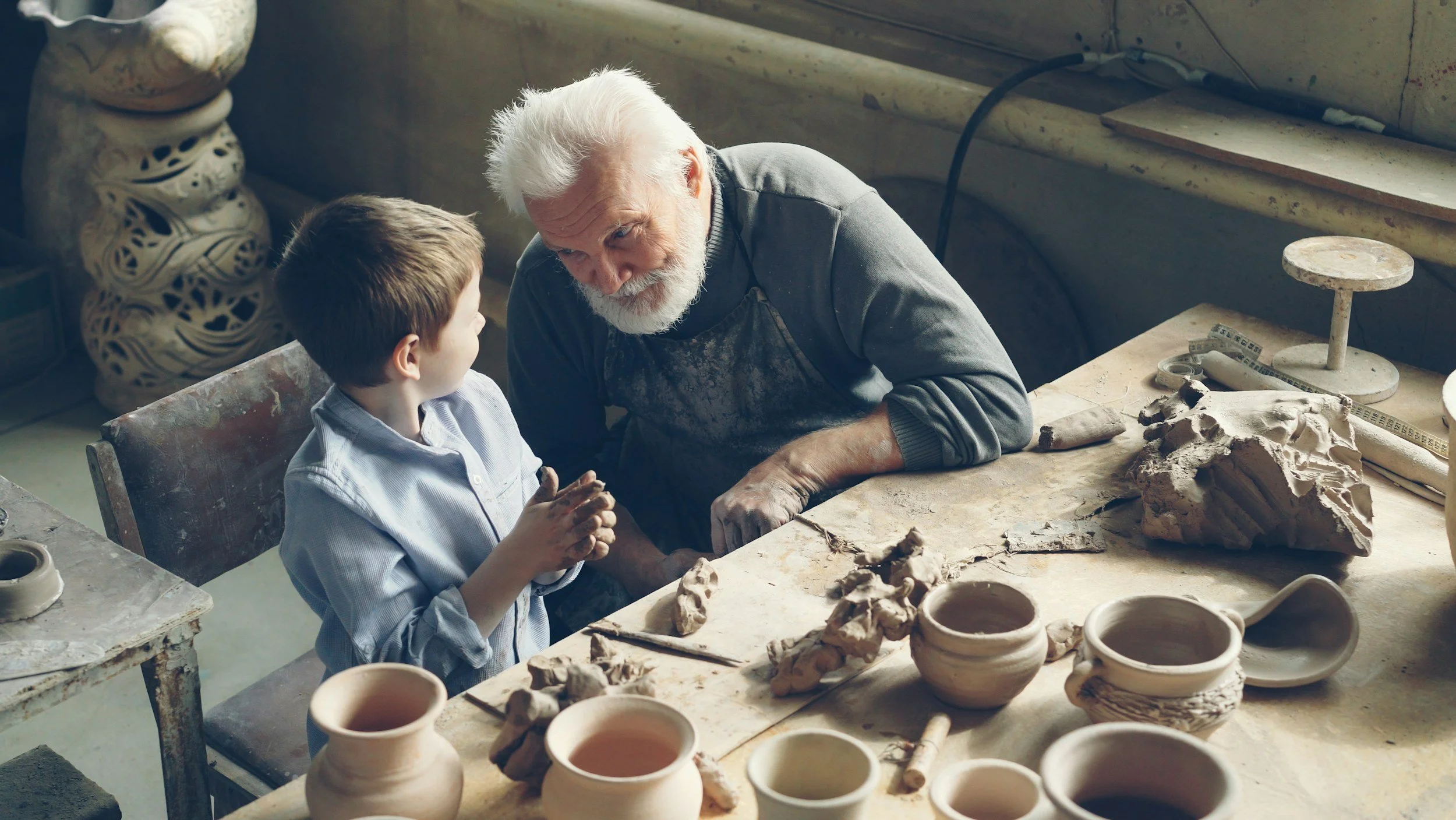 An elderly man and a young boy sitting at a pottery workbench in a ceramic studio, surrounded by clay and pottery tools, engaged in a conversation. Grandpa and grandson
