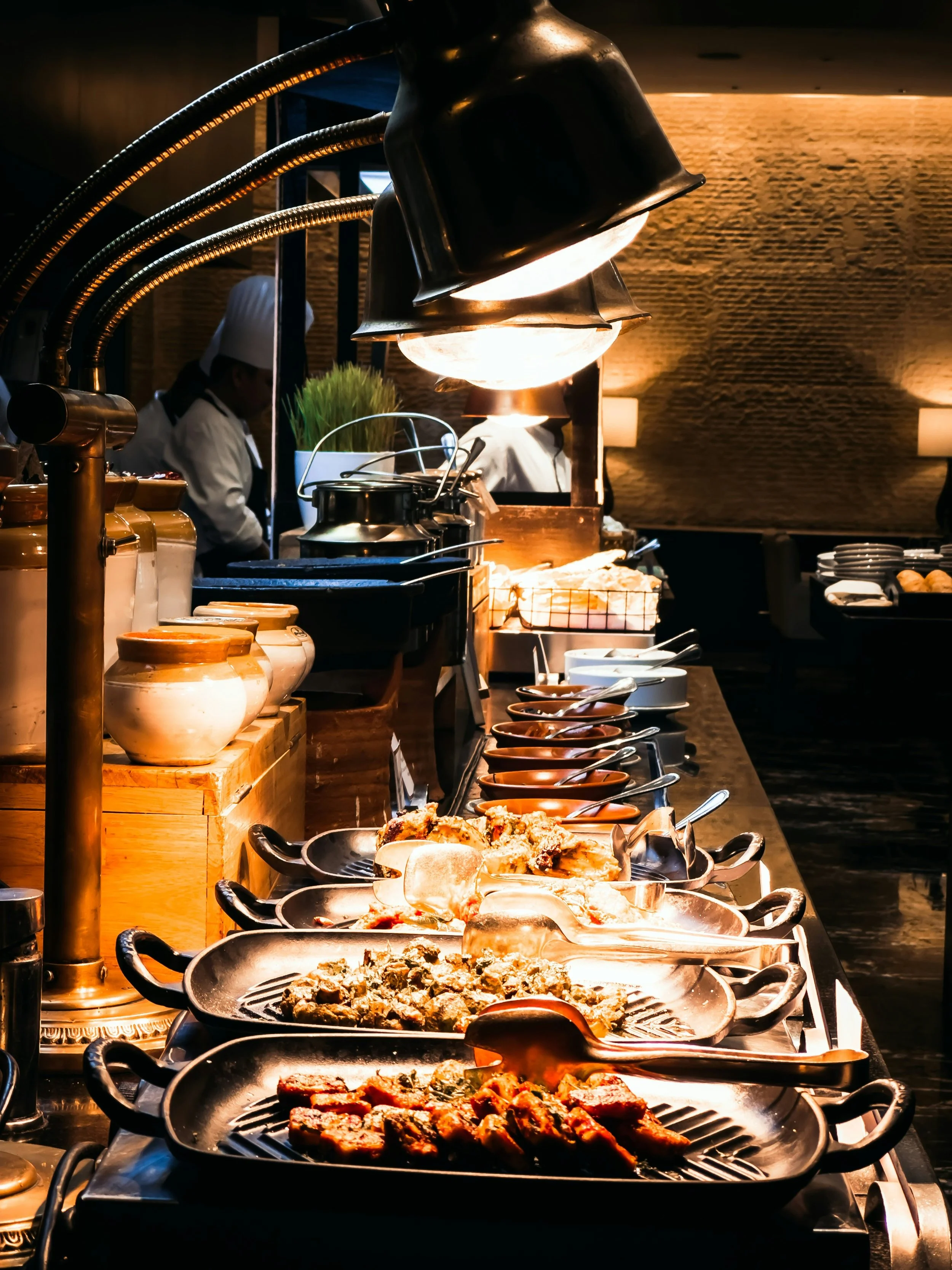 Buffet with various hot dishes in black serving trays under warm overhead lighting, with chefs preparing food in the background.