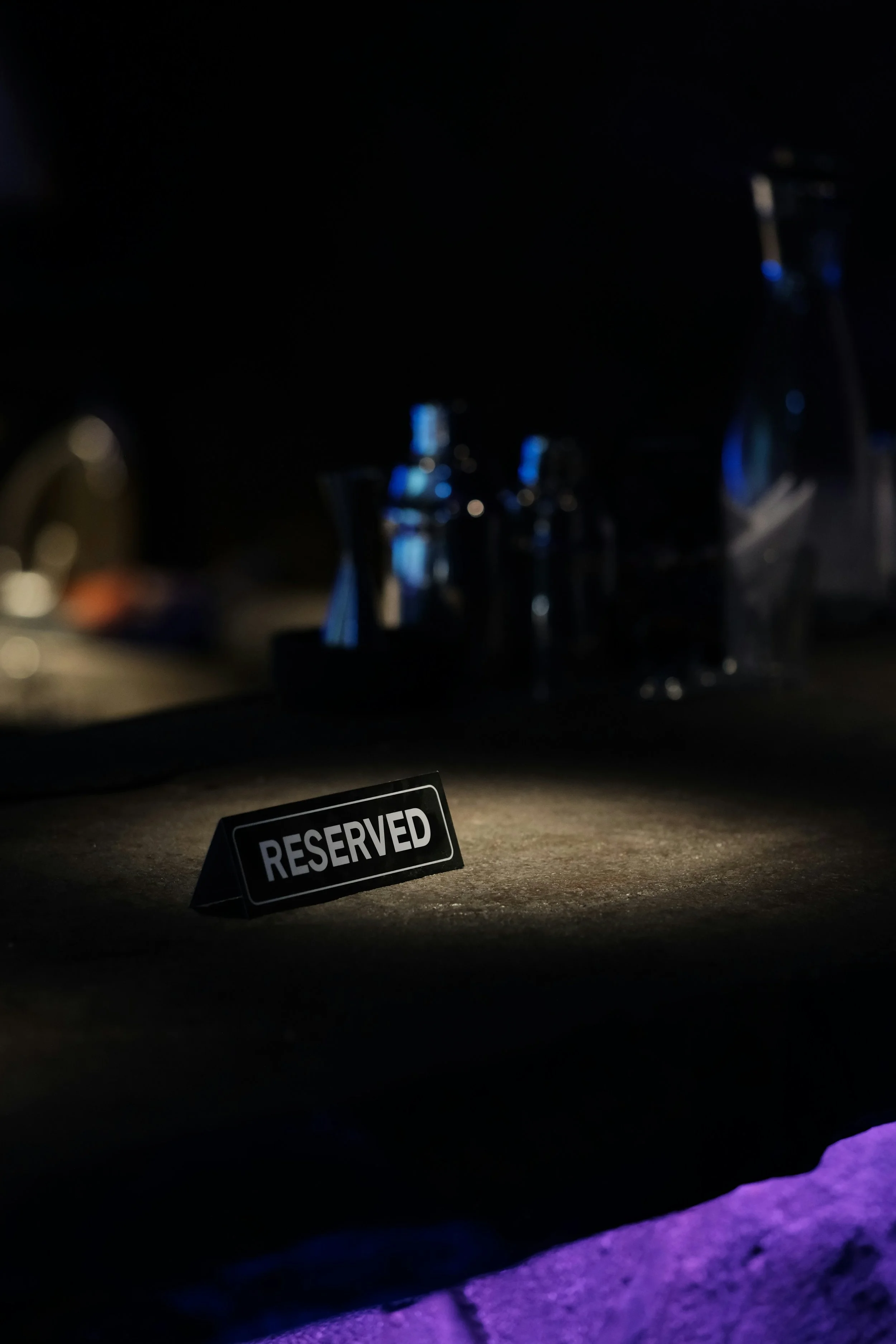 A 'Reserved' sign on a table with blurred glass bottles and pitcher in a dimly lit environment.
