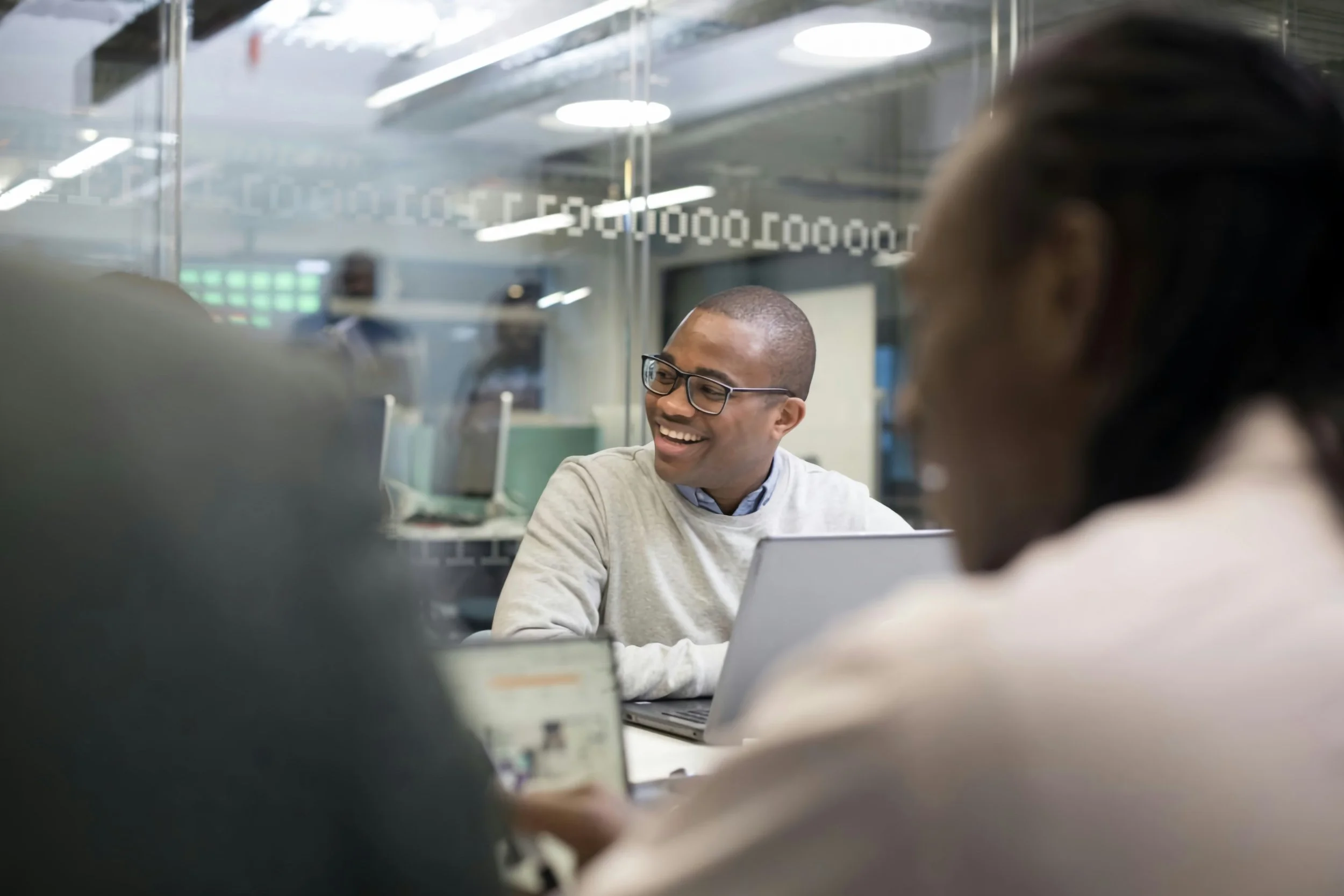 A group of diverse professionals in a meeting room, with one smiling man wearing glasses and a light gray sweater, sitting at a table with a laptop, while others surround him. The room has glass walls and modern lighting.