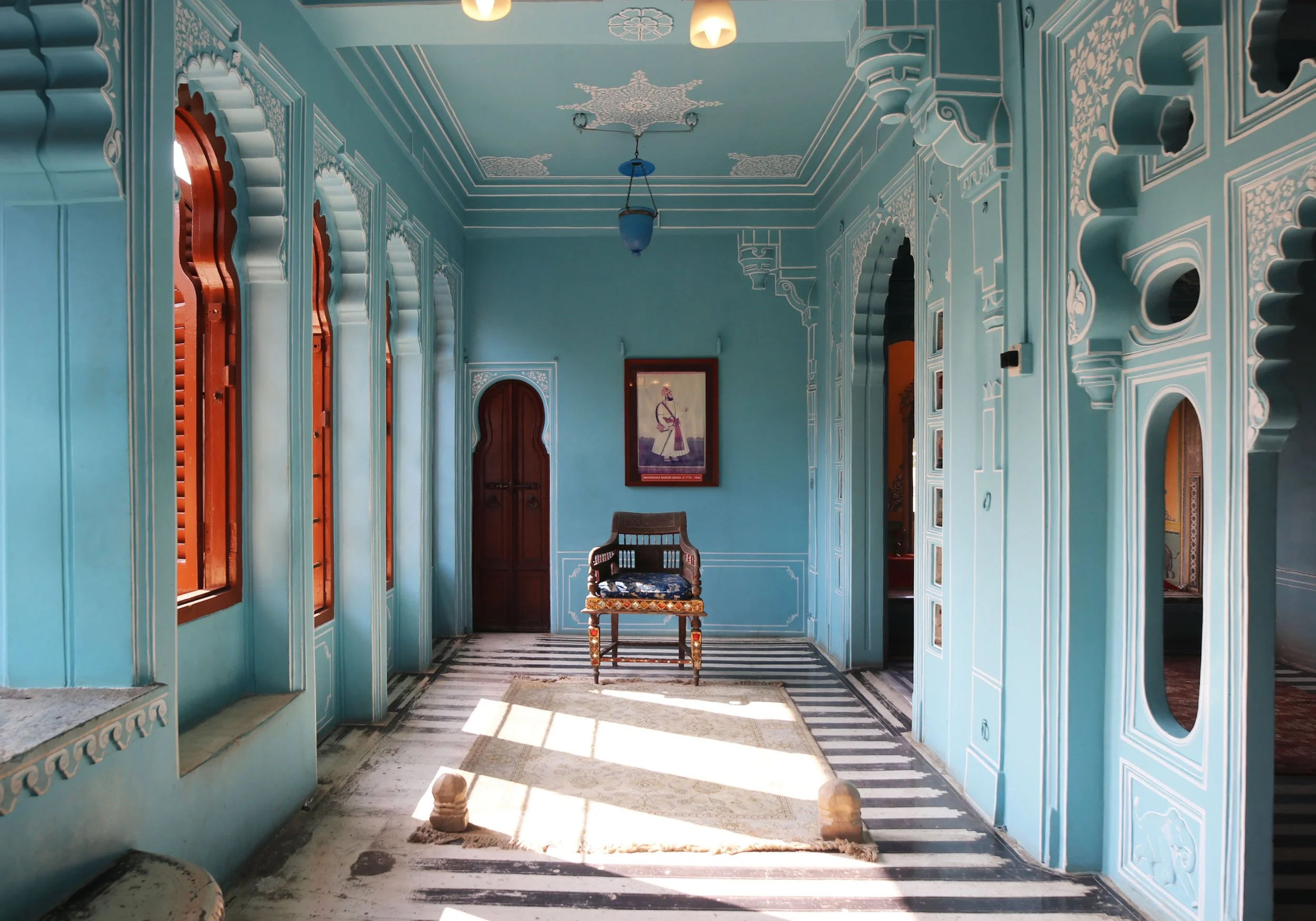 Interior of a blue room with ornate white trim, wooden doorway, and windows with wooden shutters. A framed picture hangs on the wall, and an antique wooden chair with a cushion is centered in the room. Sunlight streams in, casting shadows on a stripe