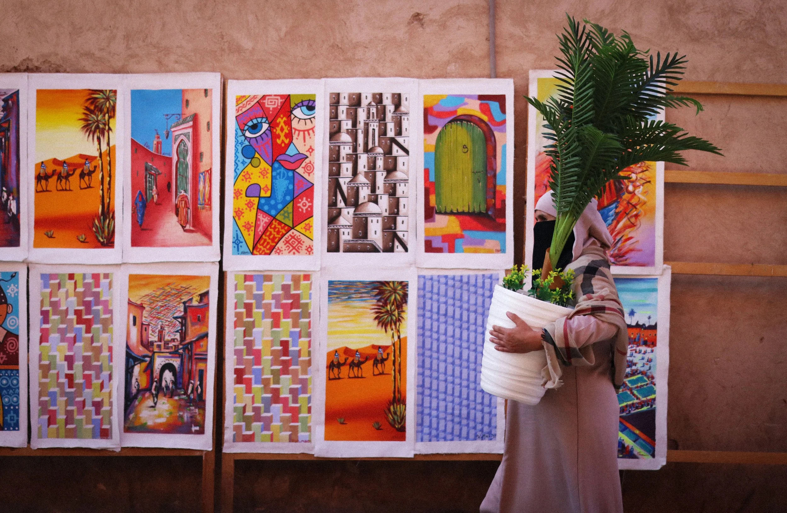A woman wearing a beige hijab and black face mask holding a white pot with a large green palm plant in front of displayed colorful artwork at an art exhibition.