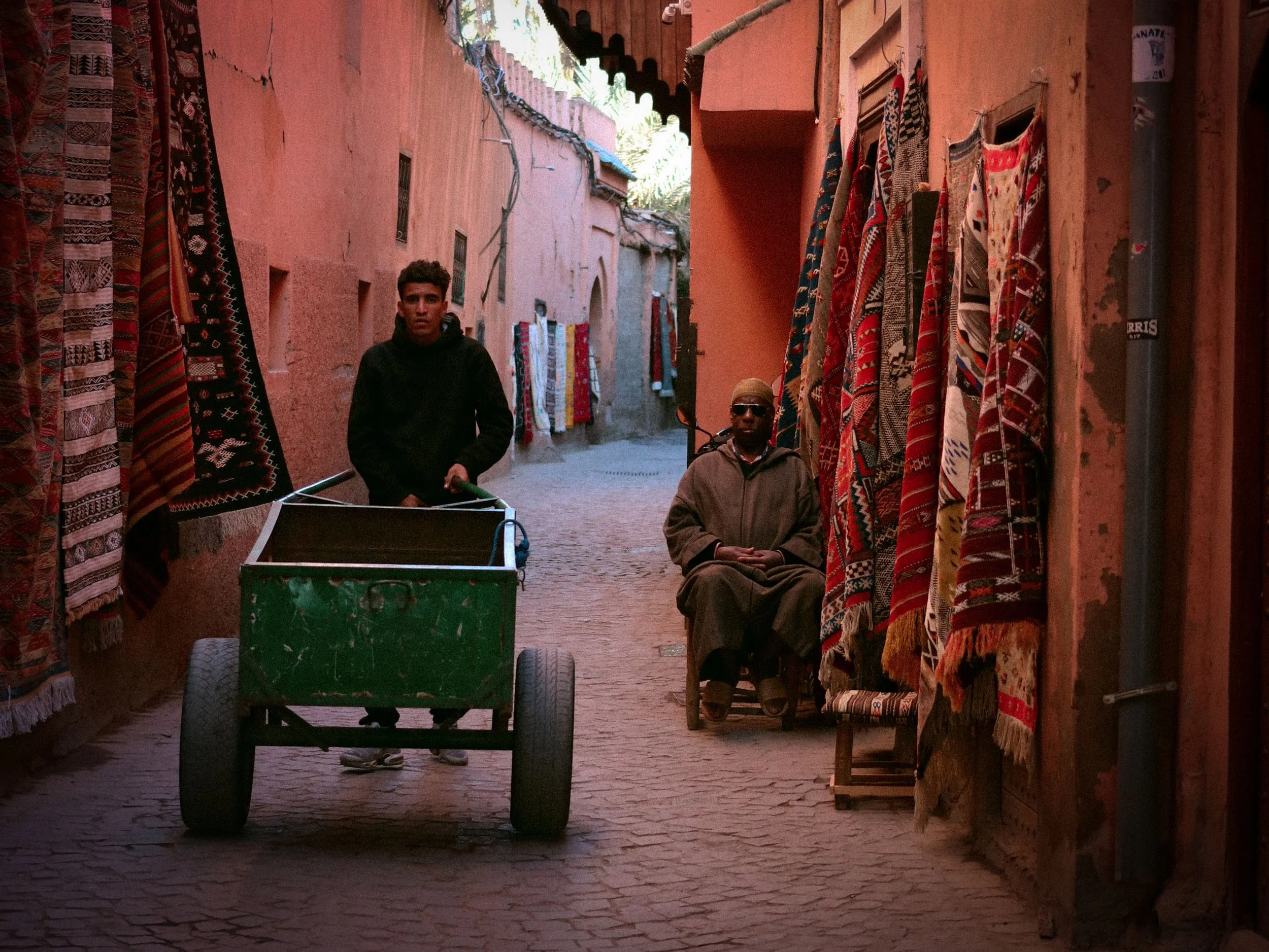 A narrow street with hanging carpets and rugs on the walls. A young man pushes a green cart, and a seated older man with sunglasses and a beige hat sits beside the wall. The street has reddish walls and cobblestone pavement.