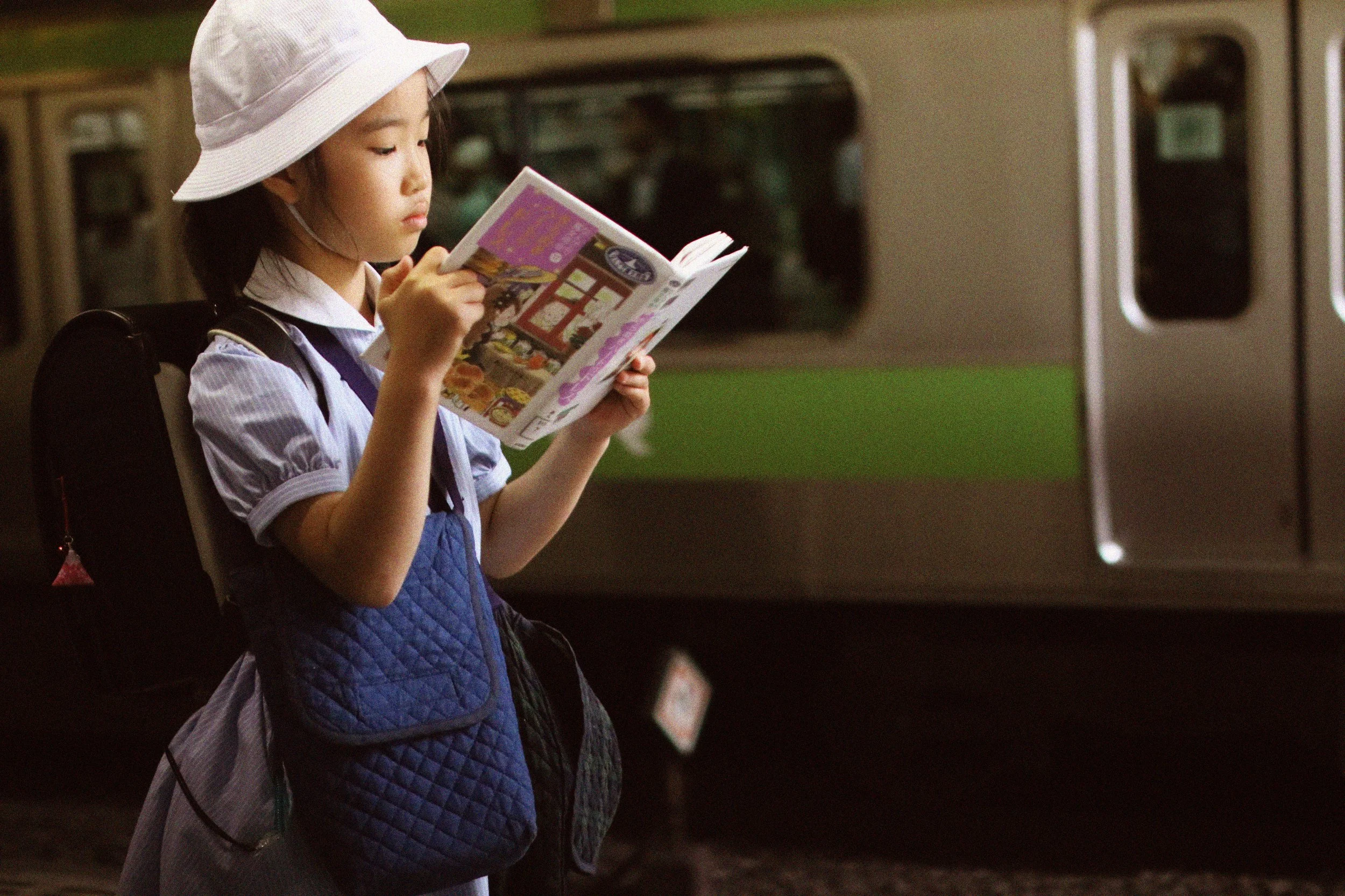 A young girl wearing a white hat and school uniform standing at a train station, reading a colorful magazine.