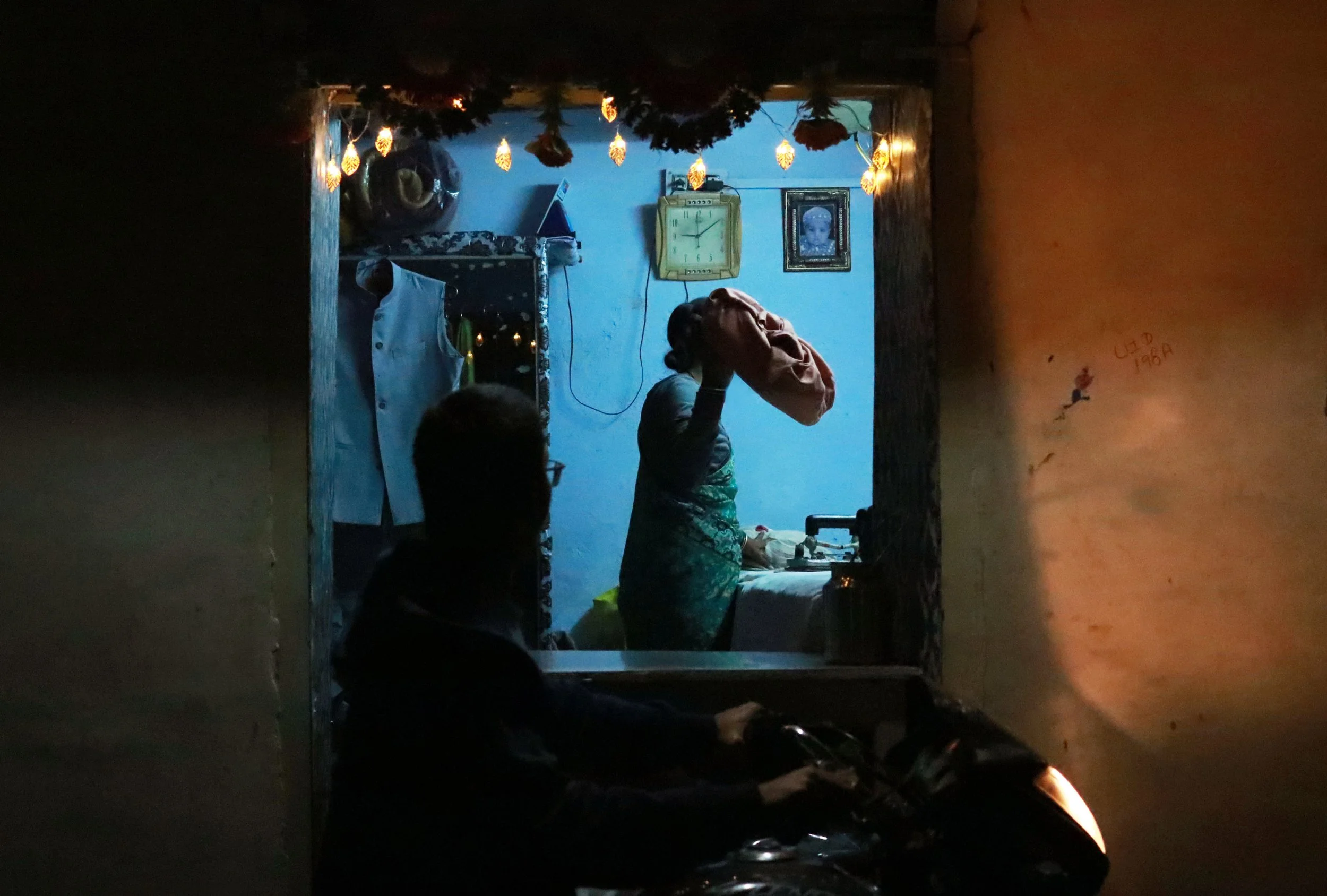 An indoor scene viewed from outside a house through a small window. Inside, a woman is lifting a large bag, and a man is riding a motorcycle passively in the foreground. The room is lit with string lights, with framed pictures, a clock, and hanging c