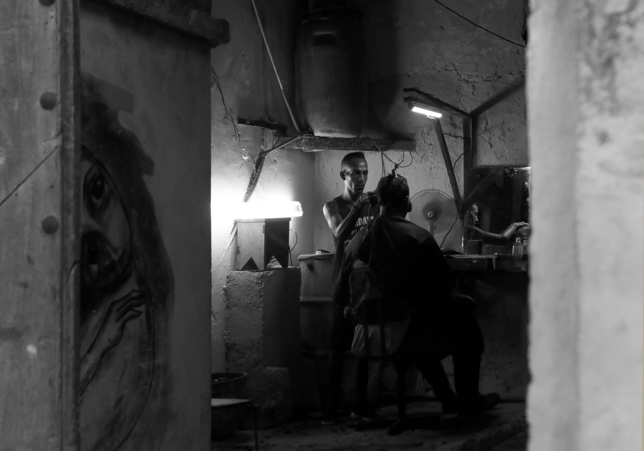 A barber cutting a person's hair in a small, dimly lit barbershop with exposed concrete walls and a mirror.