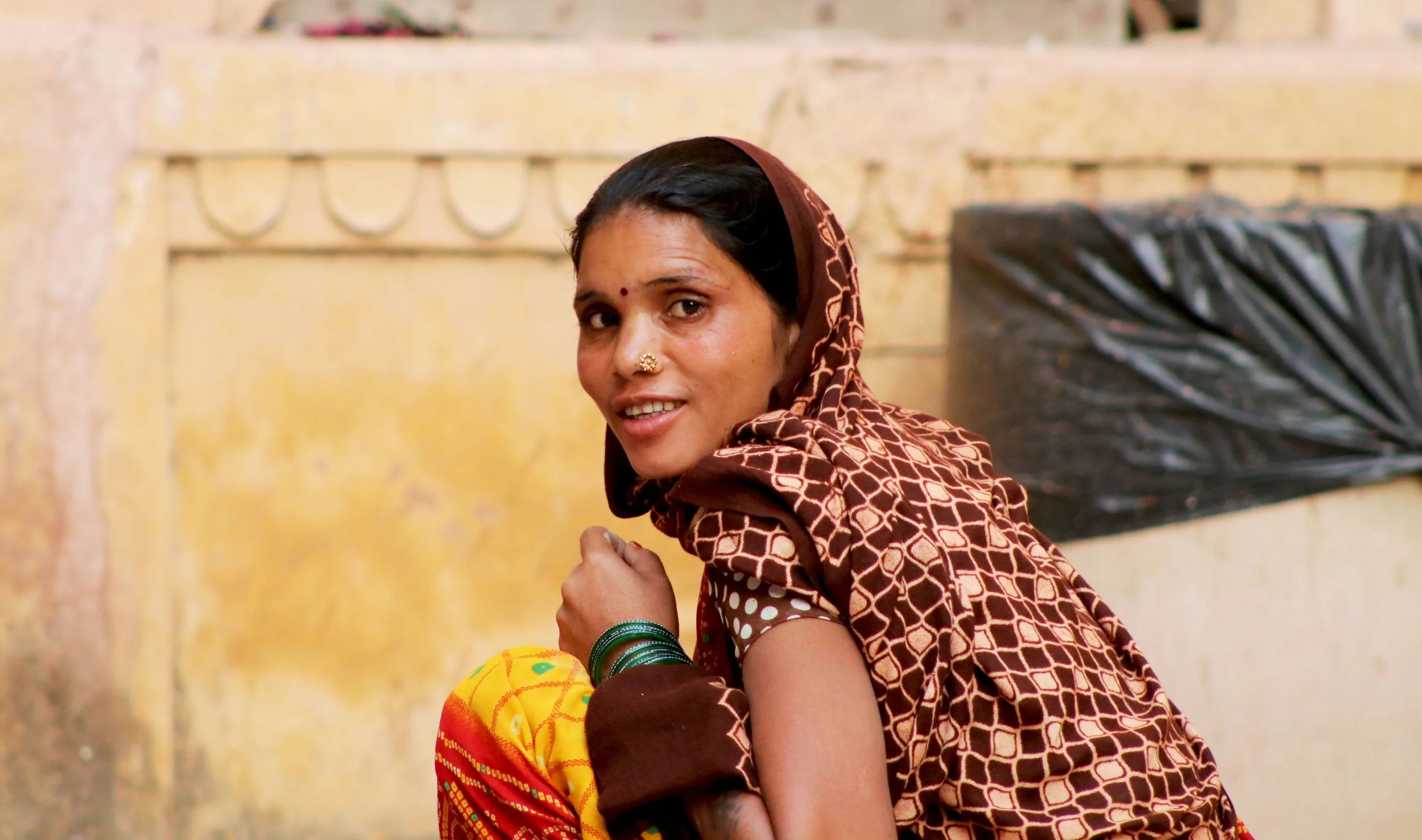 A woman sitting outdoors, wearing a patterned brown shawl and colorful clothing, with a brick wall and a black plastic-covered object in the background.