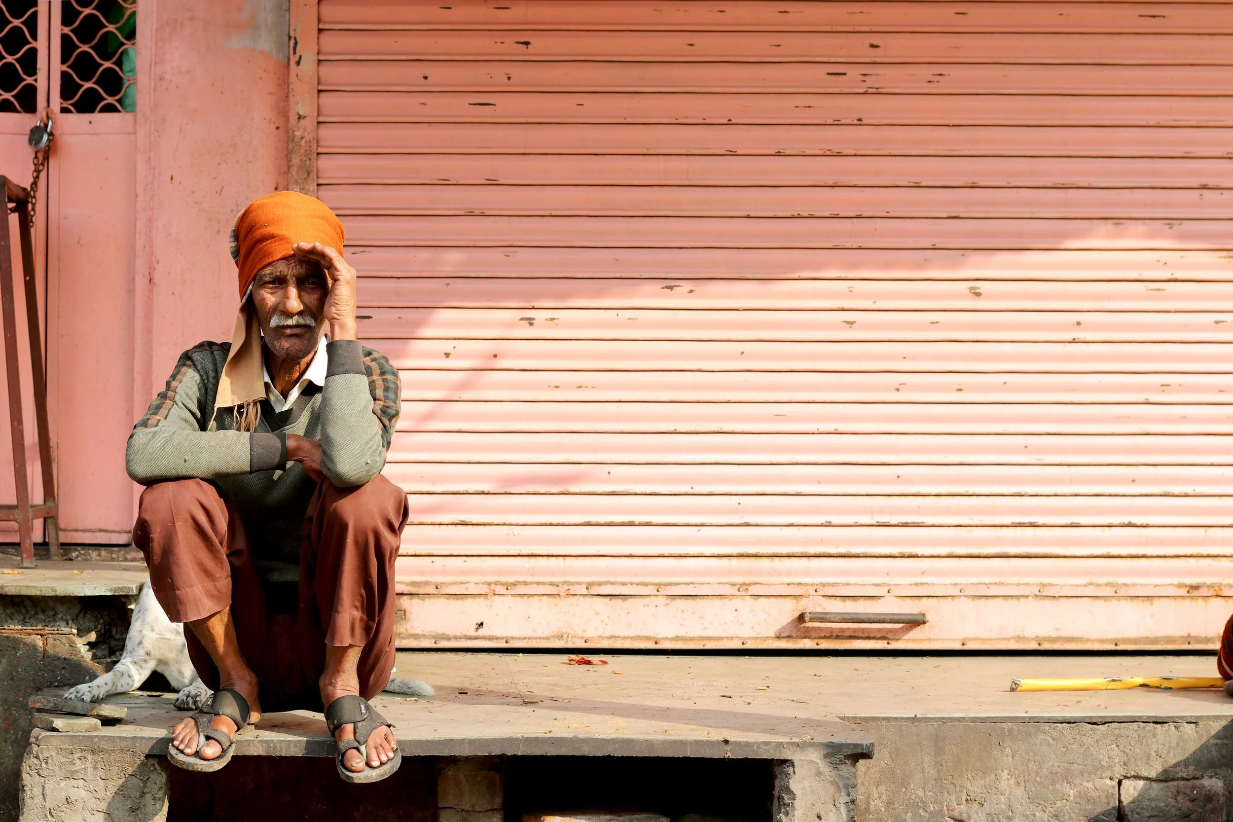 An elderly man sitting on a step in front of a pink metal shutter, wearing an orange turban, a gray sweater, brown pants, and sandals. A dog is resting behind him, partially visible.
